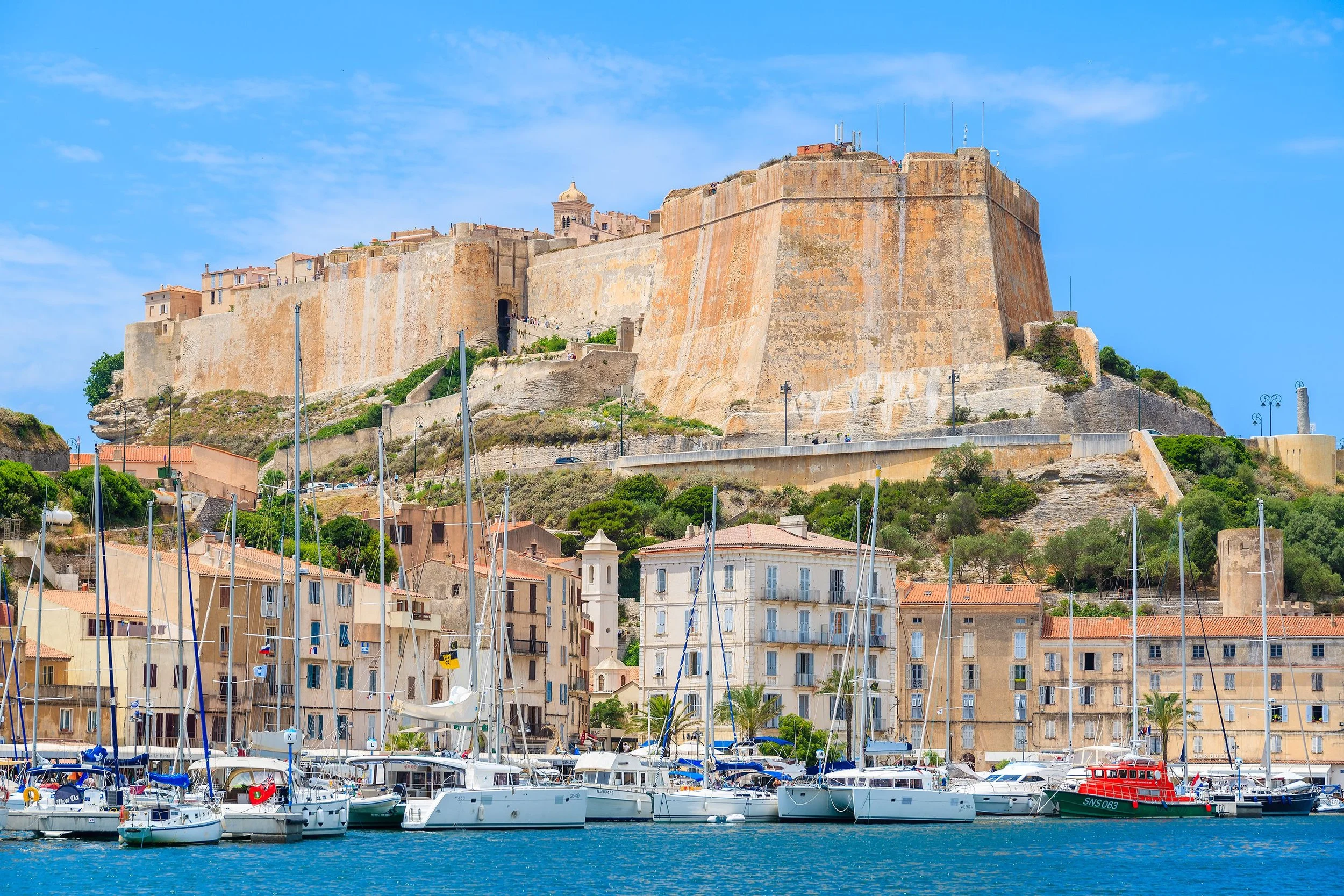 A historic castle atop a hill overlooking a marina with sailboats and yachts in a coastal town with pastel-colored buildings and green trees under a blue sky.