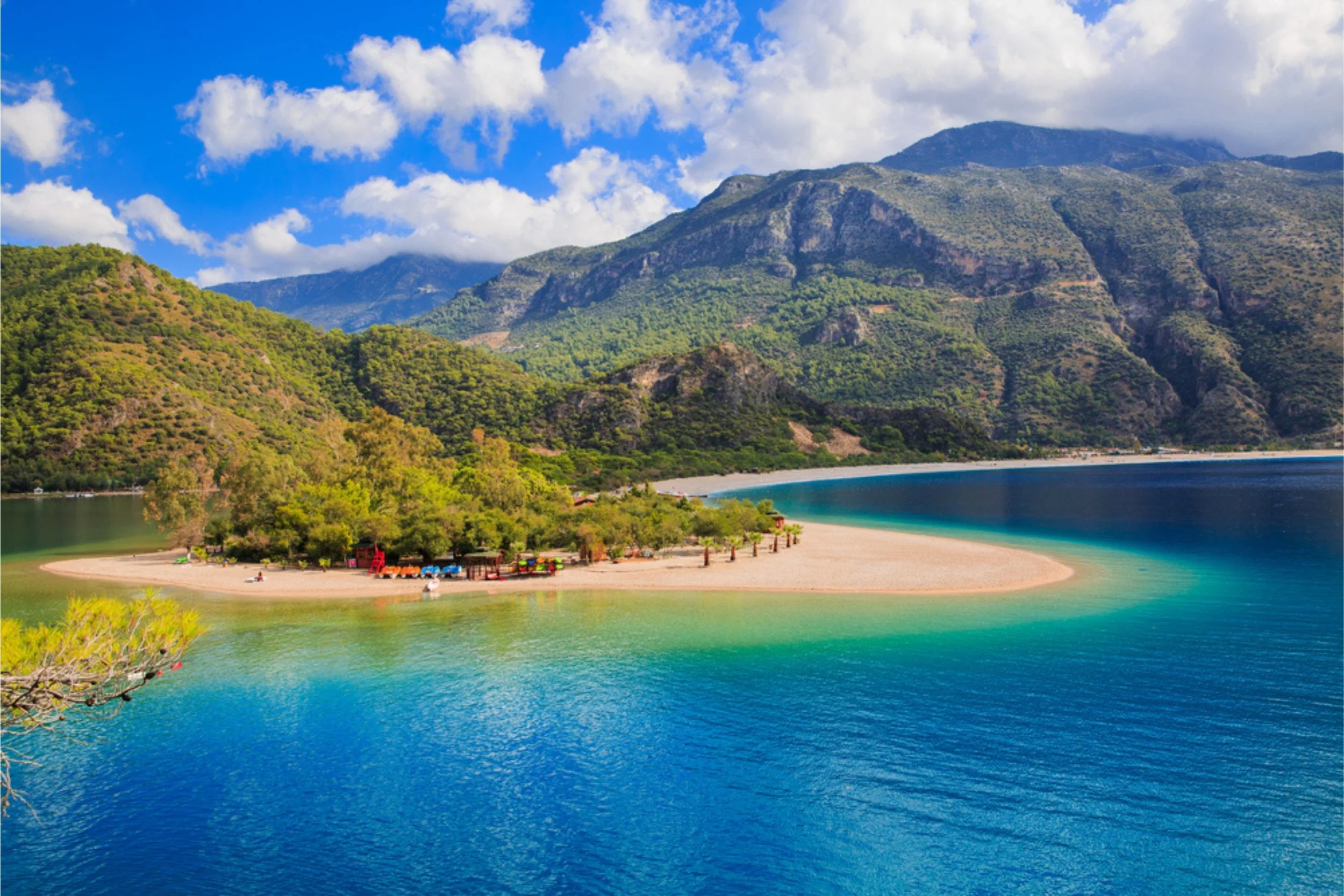 A scenic view of a curved beach cove with clear turquoise waters, surrounded by green hills and mountains under a partly cloudy sky.