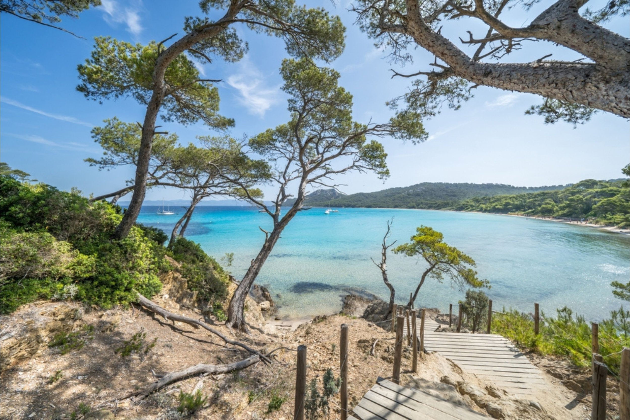 Beach with a wooden pathway leading down to the sandy shore, surrounded by trees and greenery, with blue waters and sailboats in the distance under a partly cloudy sky.