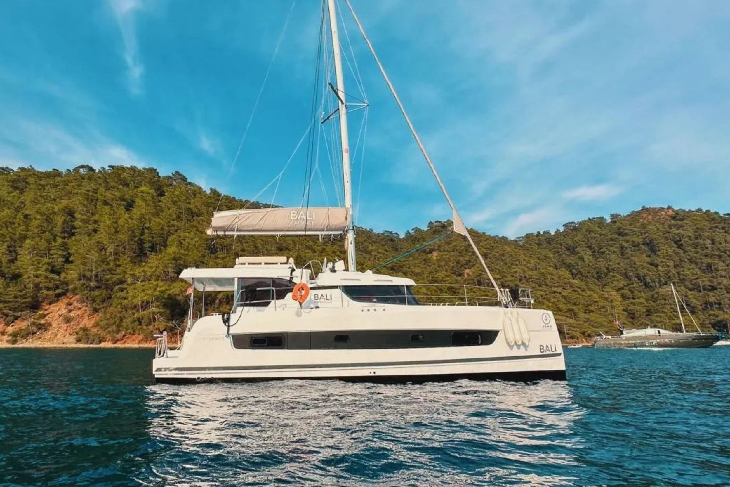 A white yacht named "BALI" sailing on the water with green hills and boats in the background under a blue sky.
