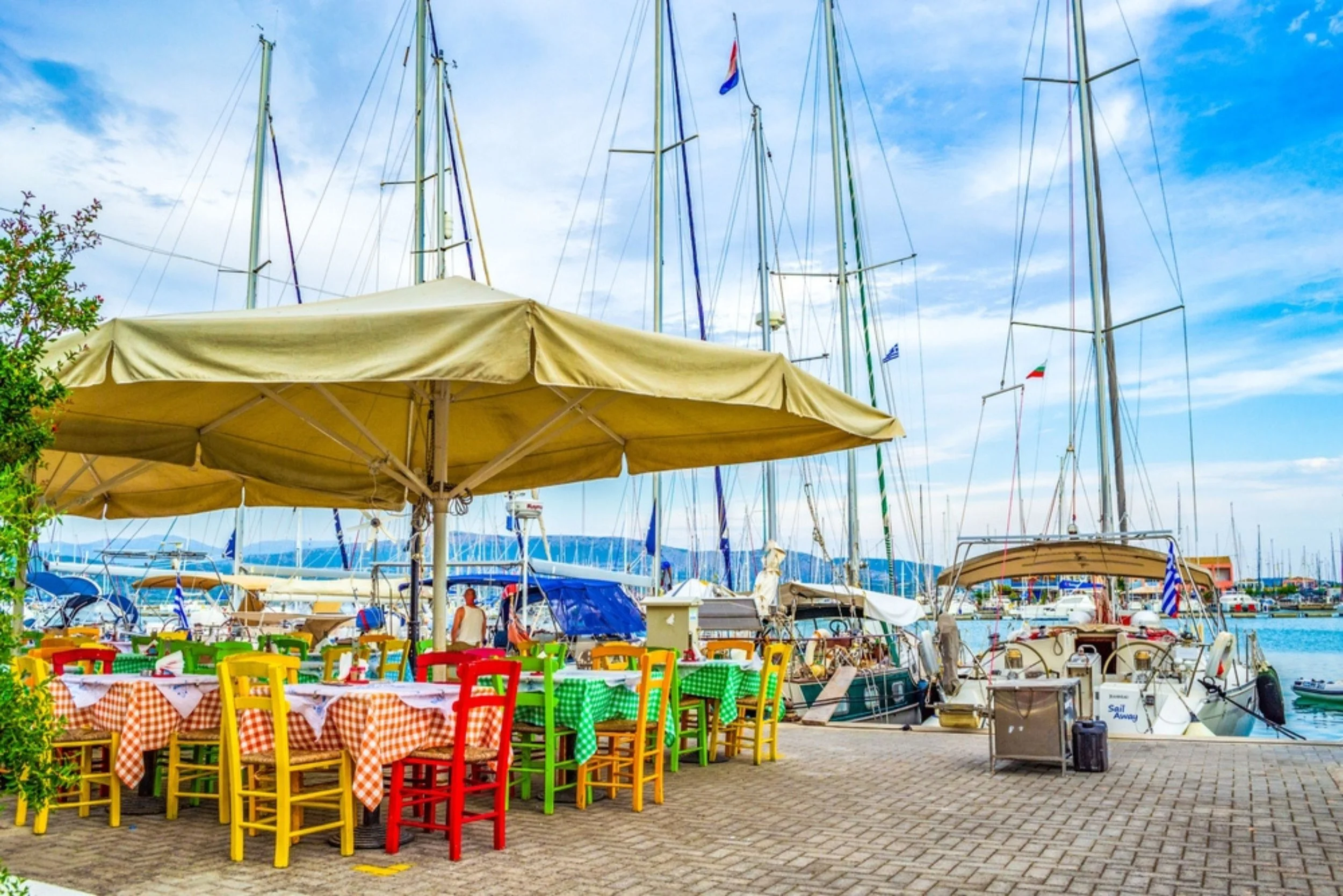 Colorful outdoor restaurant near a marina with sailboats and yachts, tables with checkered tablecloths, and a large yellow umbrella.