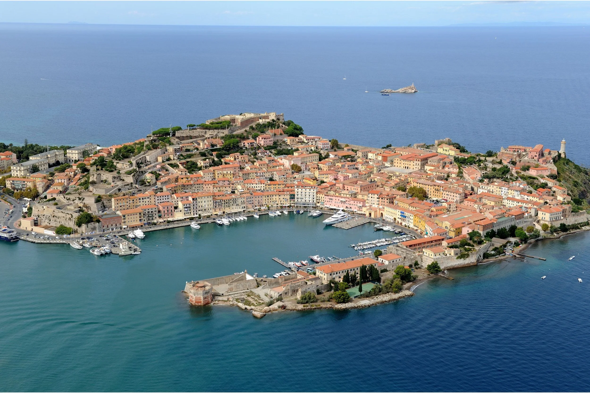 Aerial view of a historic Italian coastal town with colorful buildings, a marina filled with boats, and a fortress on a hill overlooking the sea.