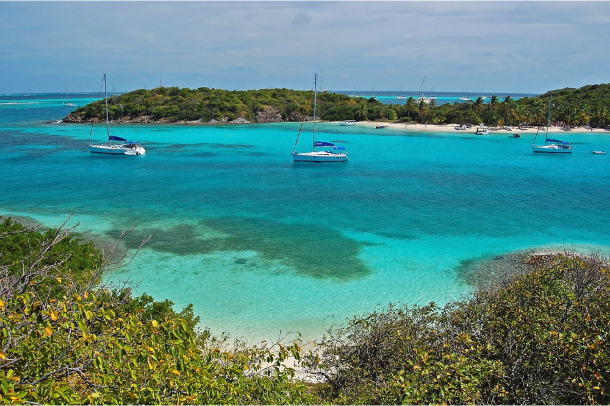 Tropical beach scene with turquoise waters, three sailboats anchored near the shore, lush green vegetation in the foreground, and a small island in the background under a partly cloudy sky.