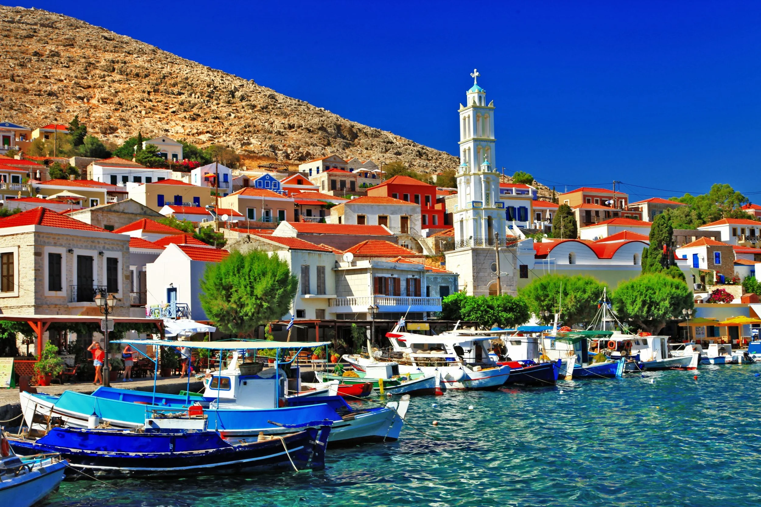 A coastal town with colorful houses, boats docked along the waterfront, and a white church with a tall steeple against a bright blue sky.