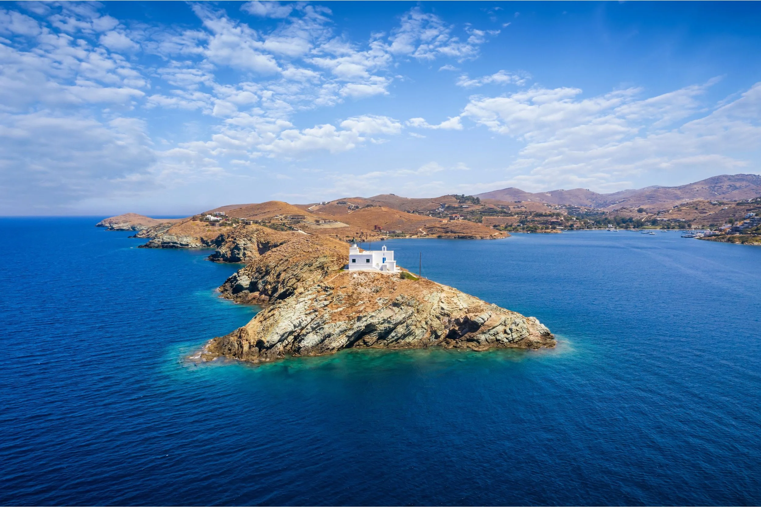 A white lighthouse on a small rocky island surrounded by blue water with a distant coastline and hills under a partly cloudy sky.