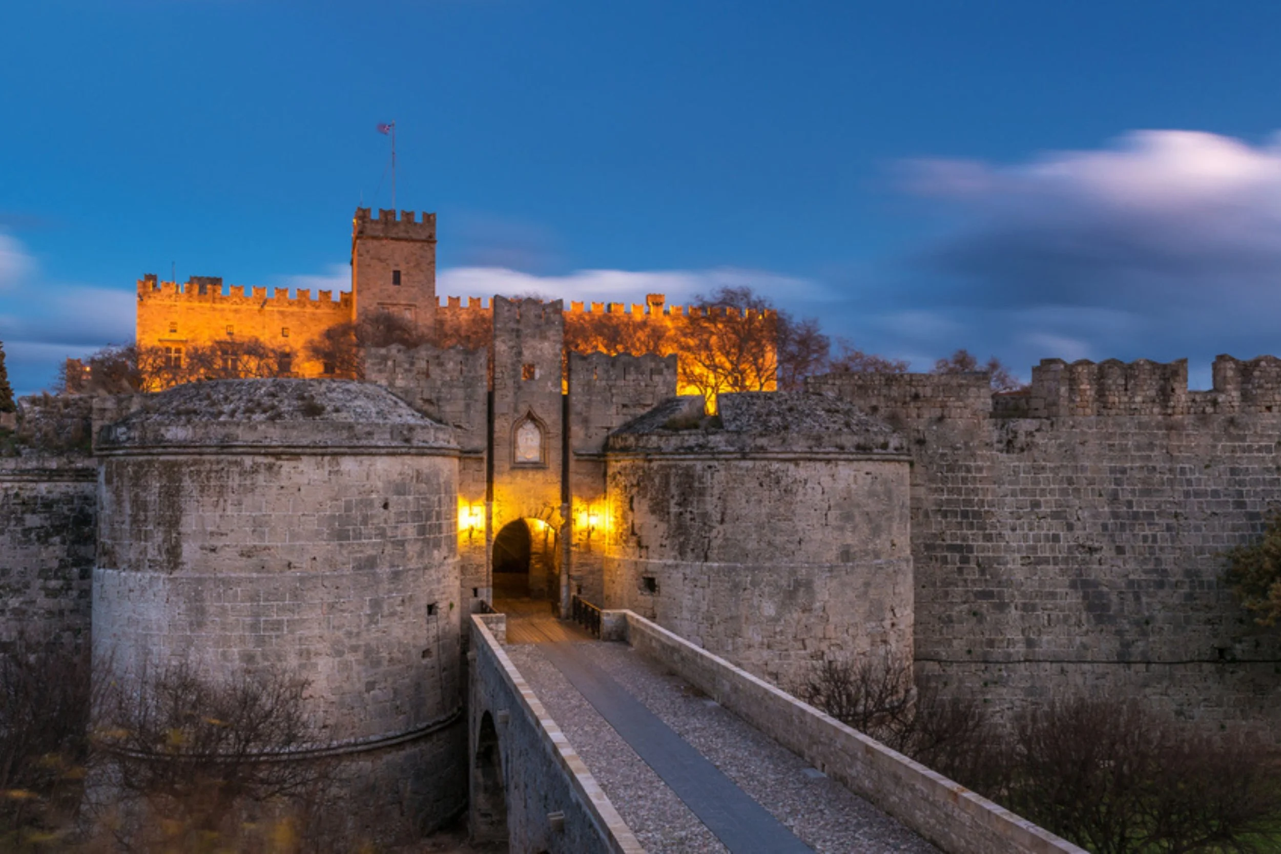Medieval stone castle with illuminated walls and a bridge at dusk, clouds in the sky.