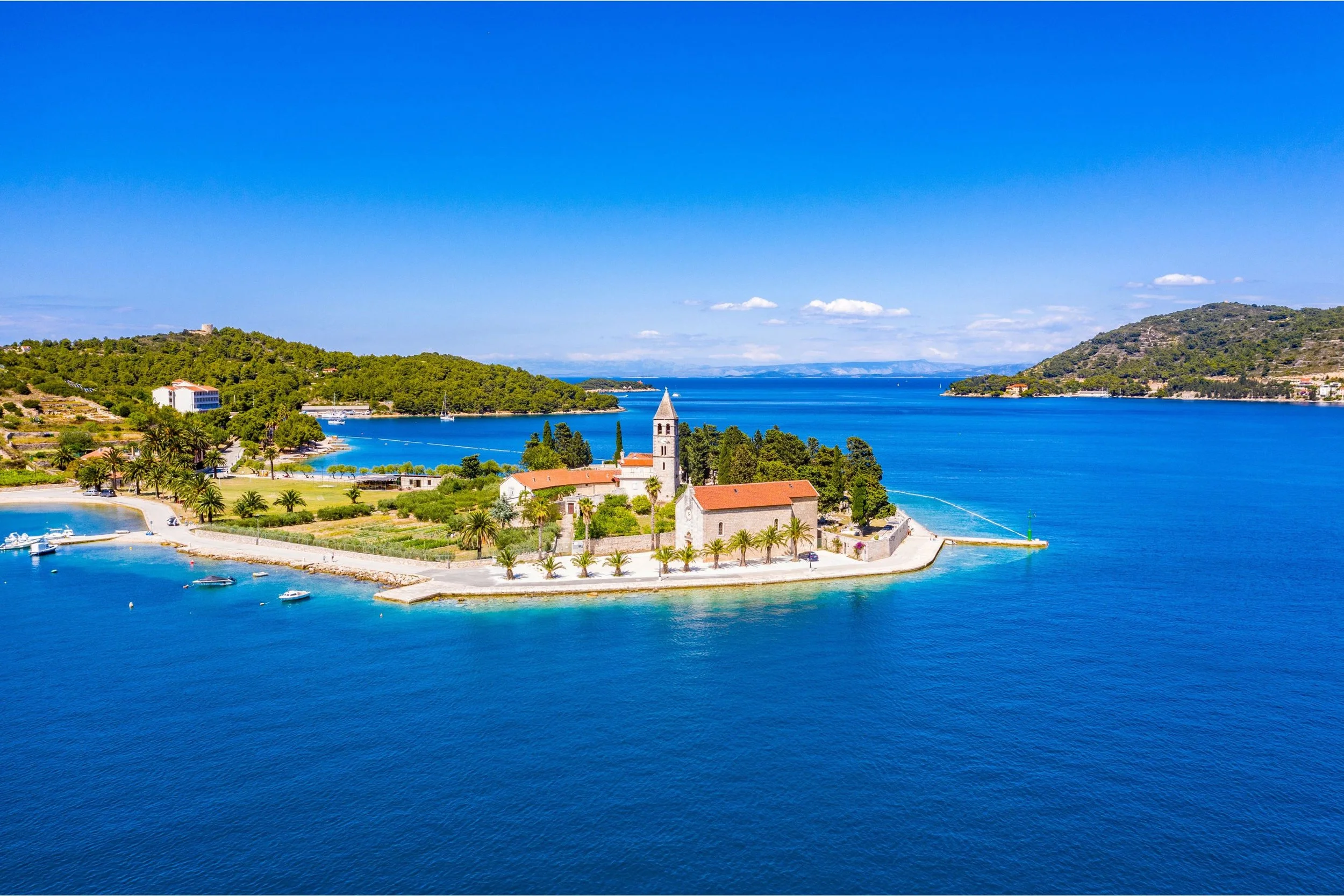 Aerial view of a small island with a church and palm trees, surrounded by blue water under a clear sky.