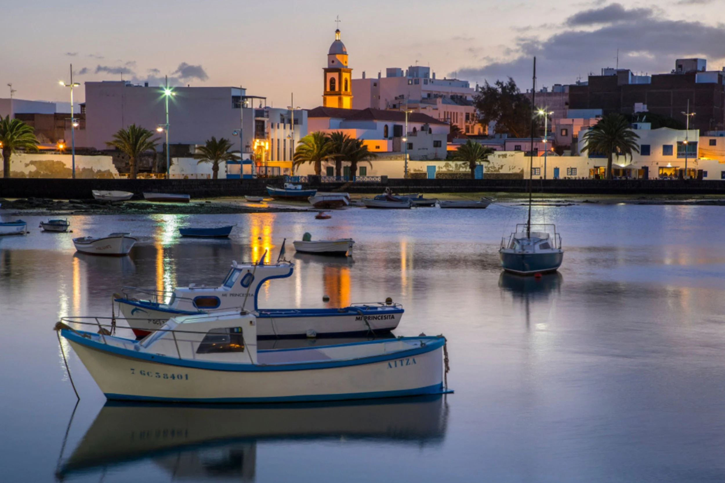 Boats moored in a calm harbor at dusk, with a town with white buildings, palm trees, and a church with a bell tower in the background, and clouds in the sky.