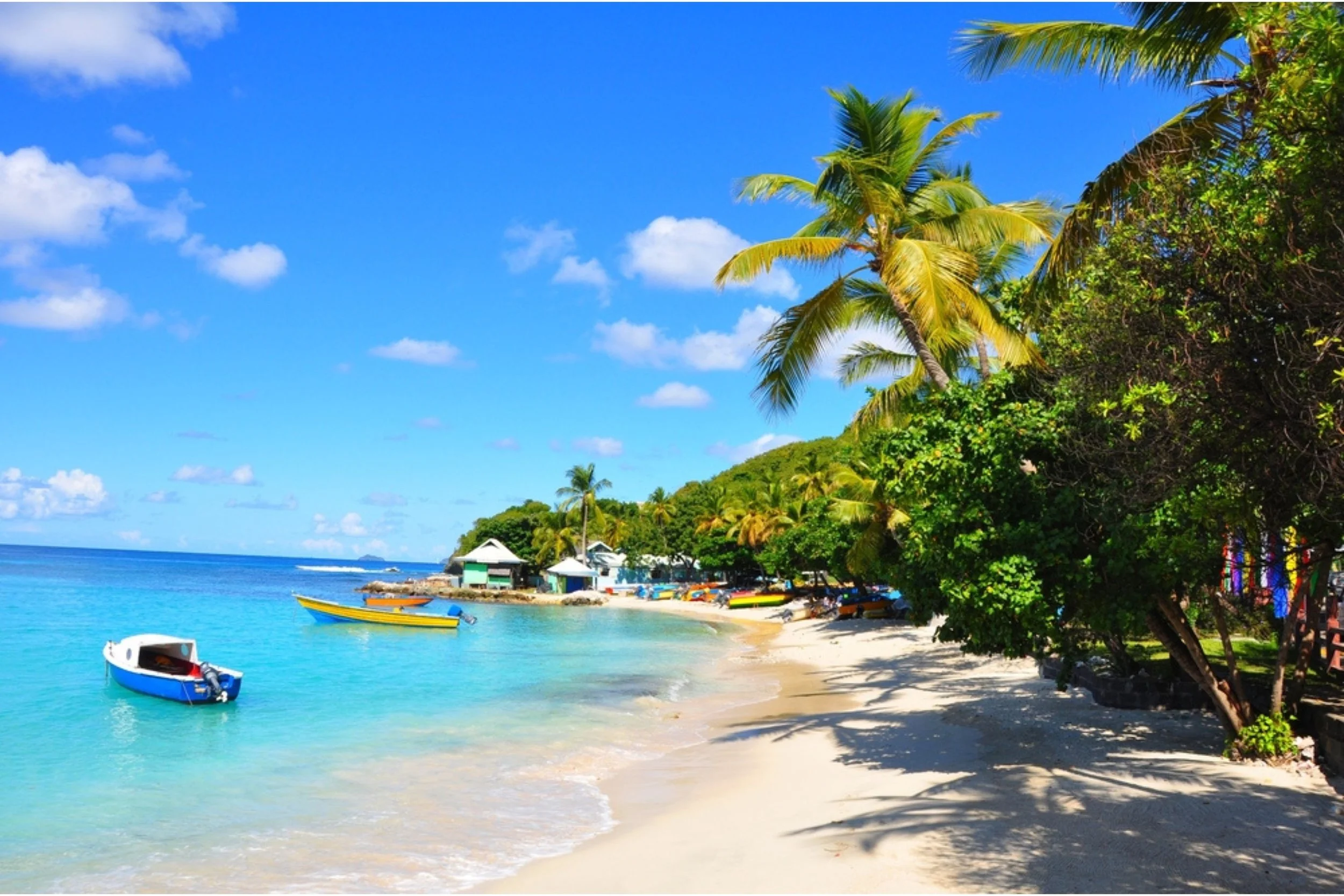 Tropical beach with clear blue water, boats, palm trees, and small beach huts under a partly cloudy sky.