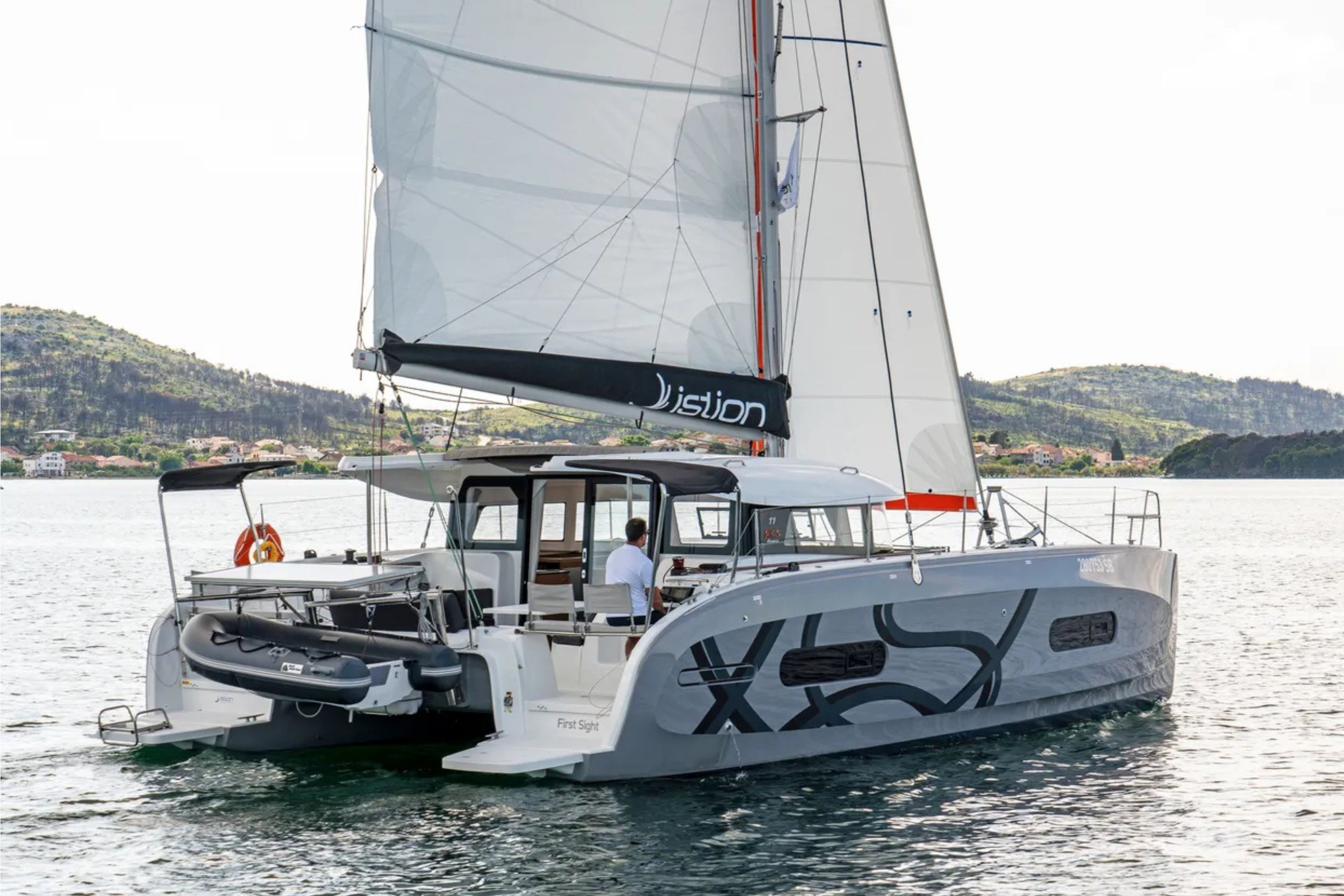 A modern sailboat with a white hull featuring black and gray abstract graphics, sailing on a body of water with mountainous shoreline in the background.