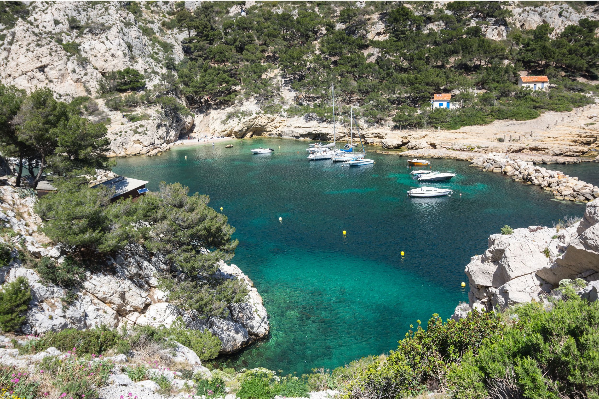 A secluded cove with clear turquoise water, surrounded by rocky cliffs and green trees. Several boats are docked along the shoreline, with a small stone jetty. Two small houses with white walls and orange roofs are visible on the hill in the background.