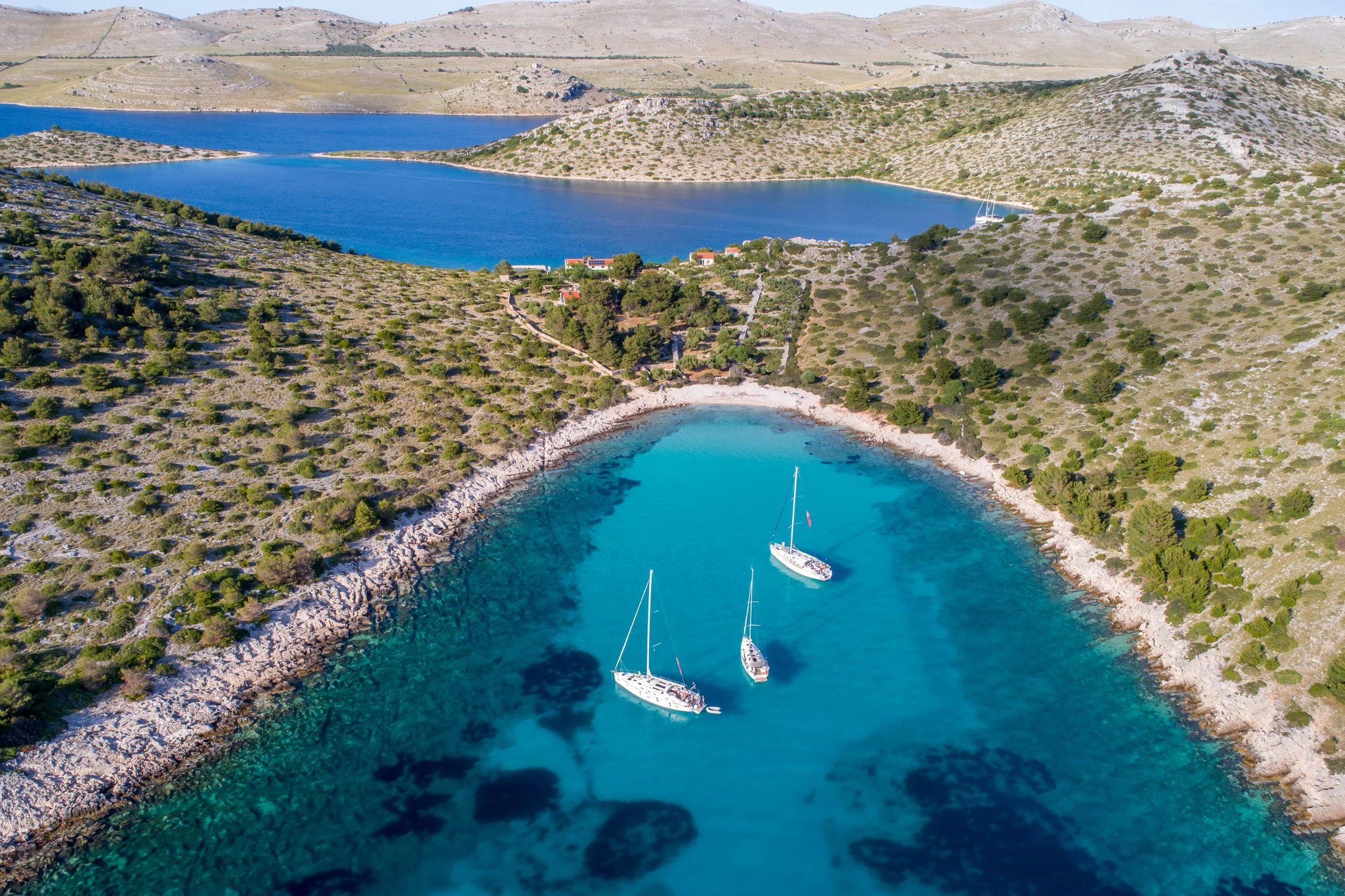 Aerial view of a small, curved cove with turquoise water, surrounded by rocky, shrub-covered hills. Three sailboats are anchored in the cove. In the background, a larger body of water and hilly terrain are visible.