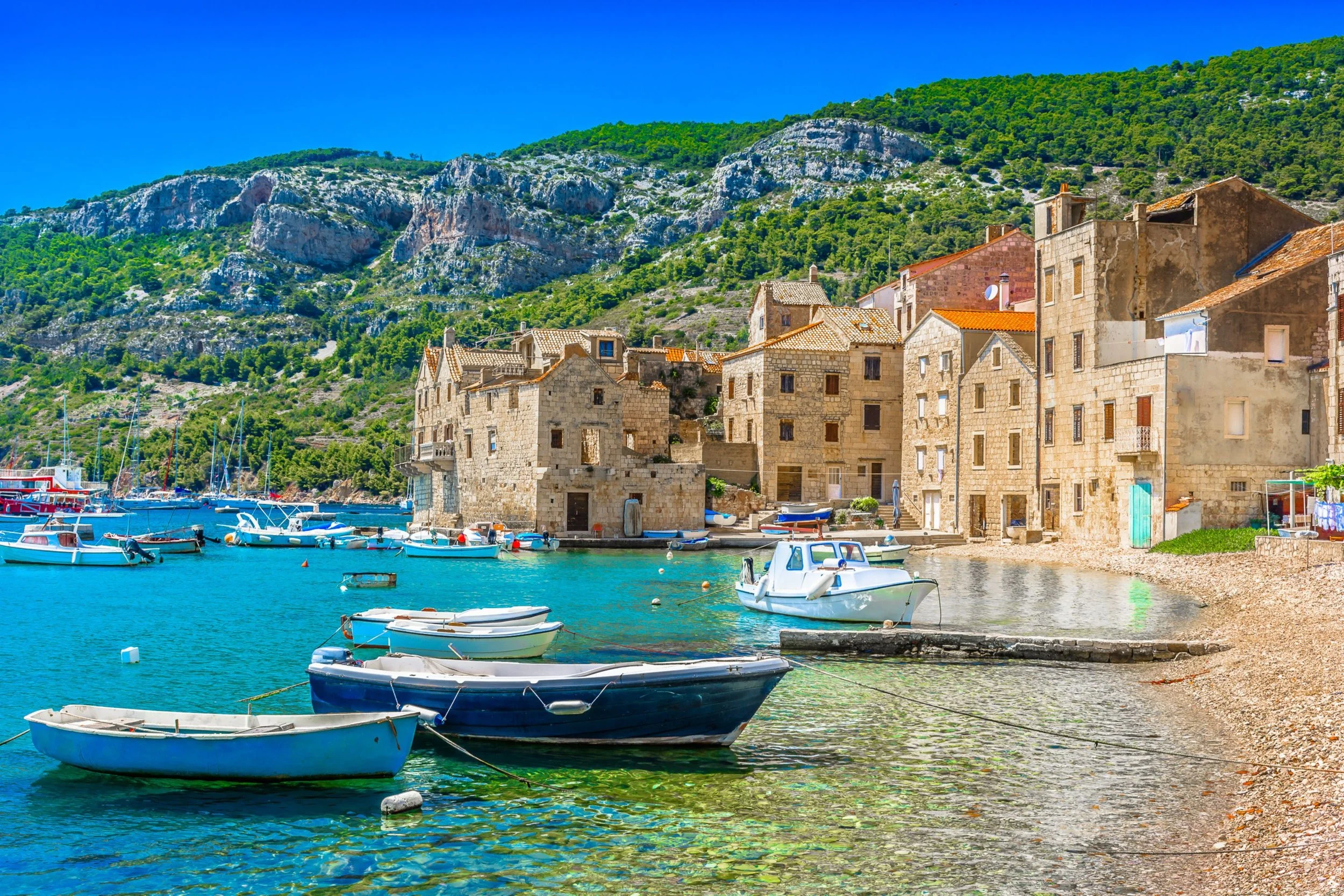 A coastal scene with historic stone buildings along the shoreline, boats docked in clear blue water, and green hills in the background under a bright blue sky.