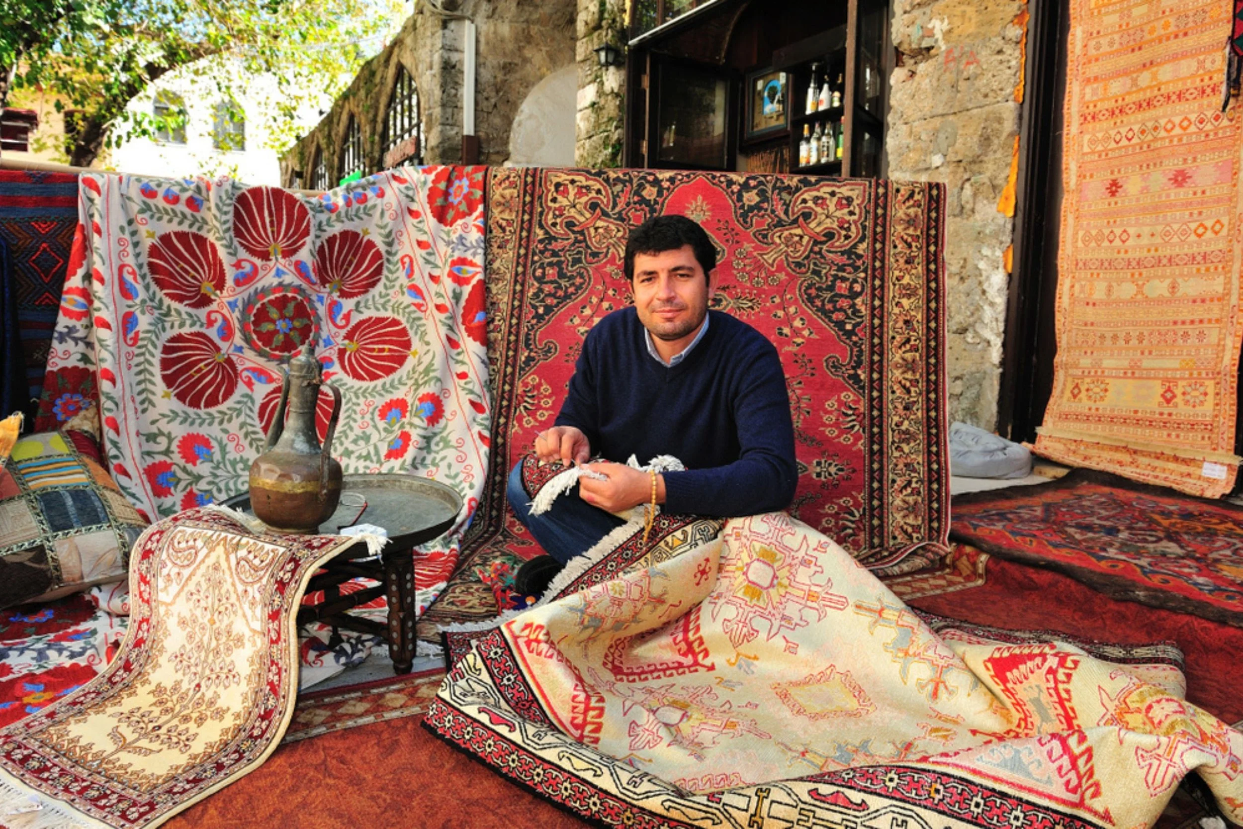 A man sitting among a display of colorful traditional rugs and textiles outdoors.