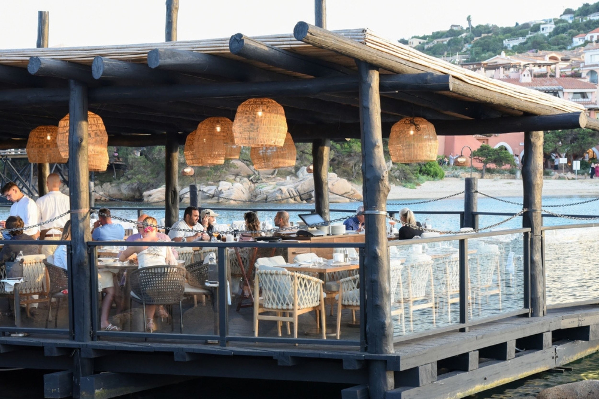 A seaside restaurant with a wooden deck, outdoor seating, hanging wicker lamps, and a view of a beach and hillside houses.