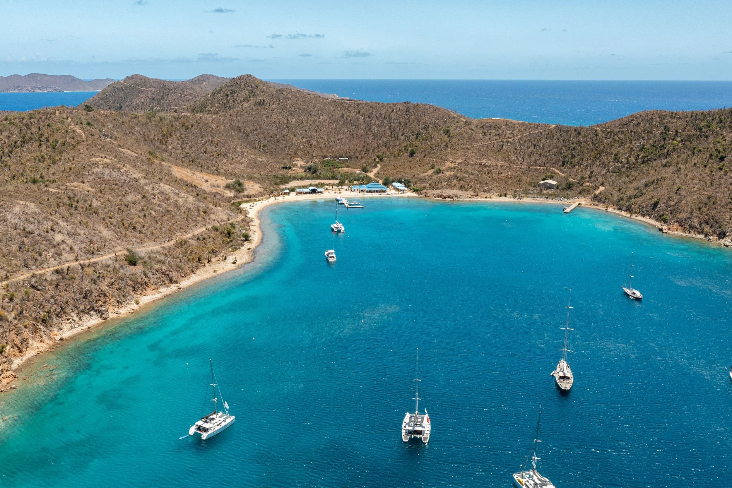 Aerial view of a scenic bay with clear blue water, surrounded by brown hills. Several sailboats are anchored in the water near a small sandy beach, with some buildings and pathways visible on the shoreline.