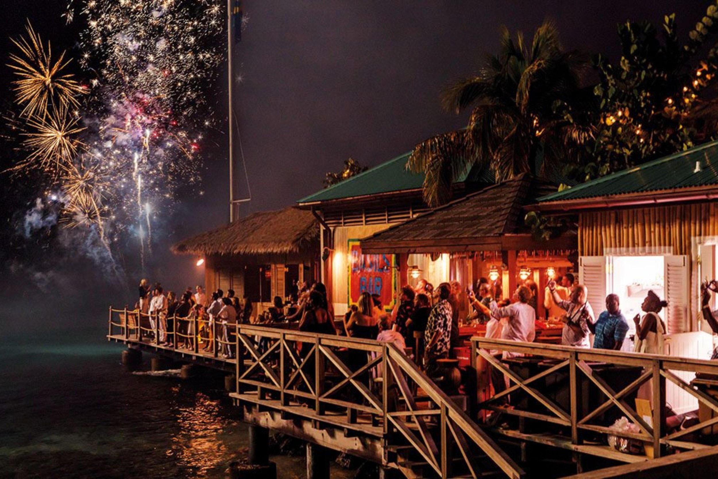 People gathered on a wooden deck by the water watching fireworks at night, with a building illuminated inside and palm trees in the background.