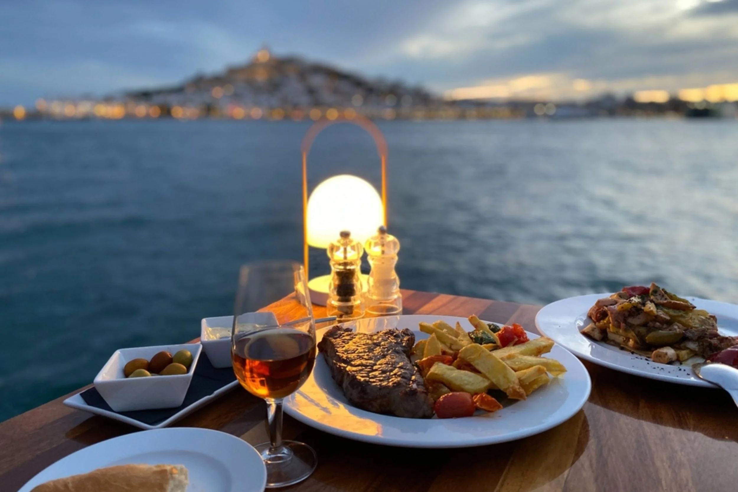 Dinner table set with steak, fries, vegetables, a glass of wine, olives, and a bowl of sauce, overlooking a body of water near a coastal town at sunset.