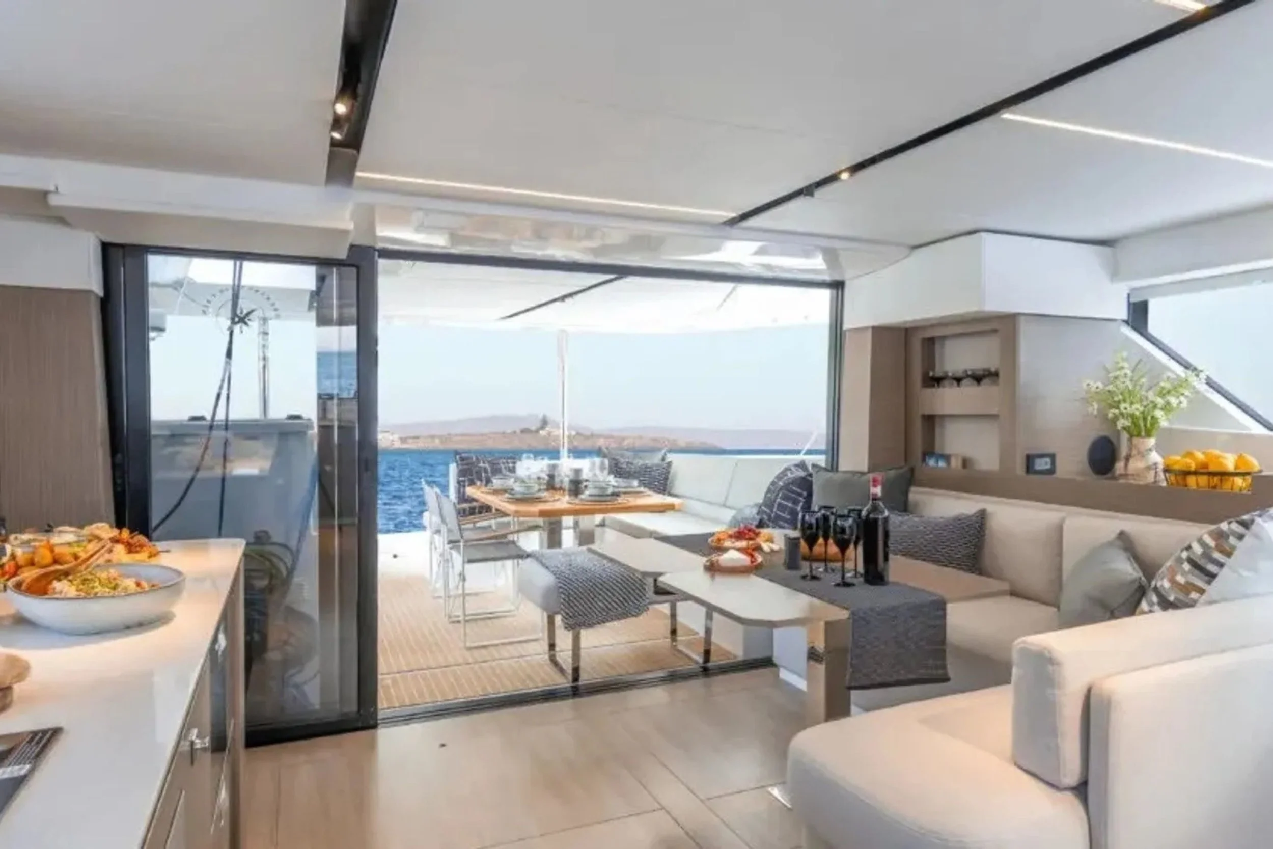 Interior of a yacht living area with a view of water and sky outside, decorated with a beige sectional sofa, a coffee table, and a dining table set for a meal on a balcony.
