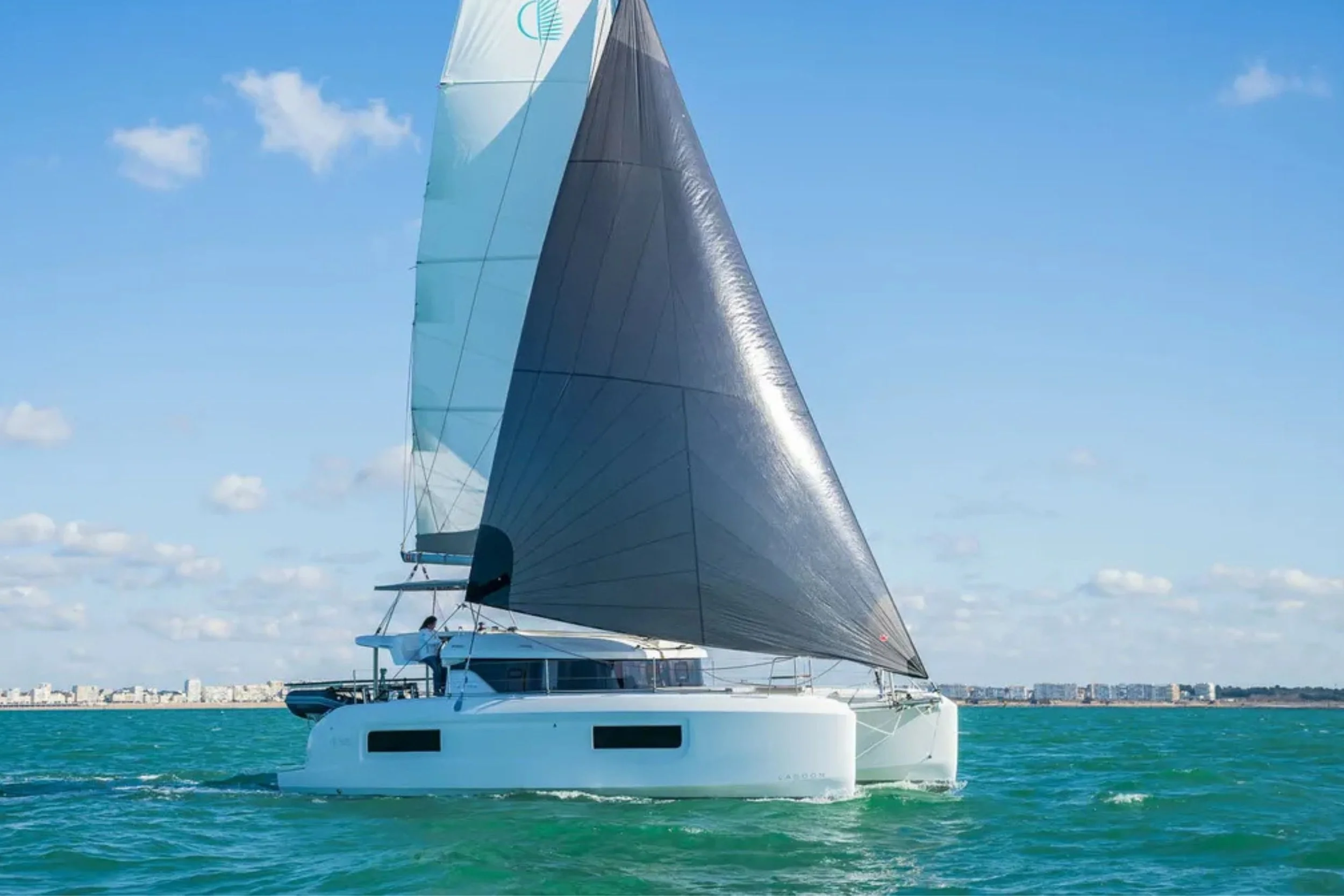 A large modern sailboat with black sails and a white hull sailing on turquoise water on a sunny day with a city skyline in the background.