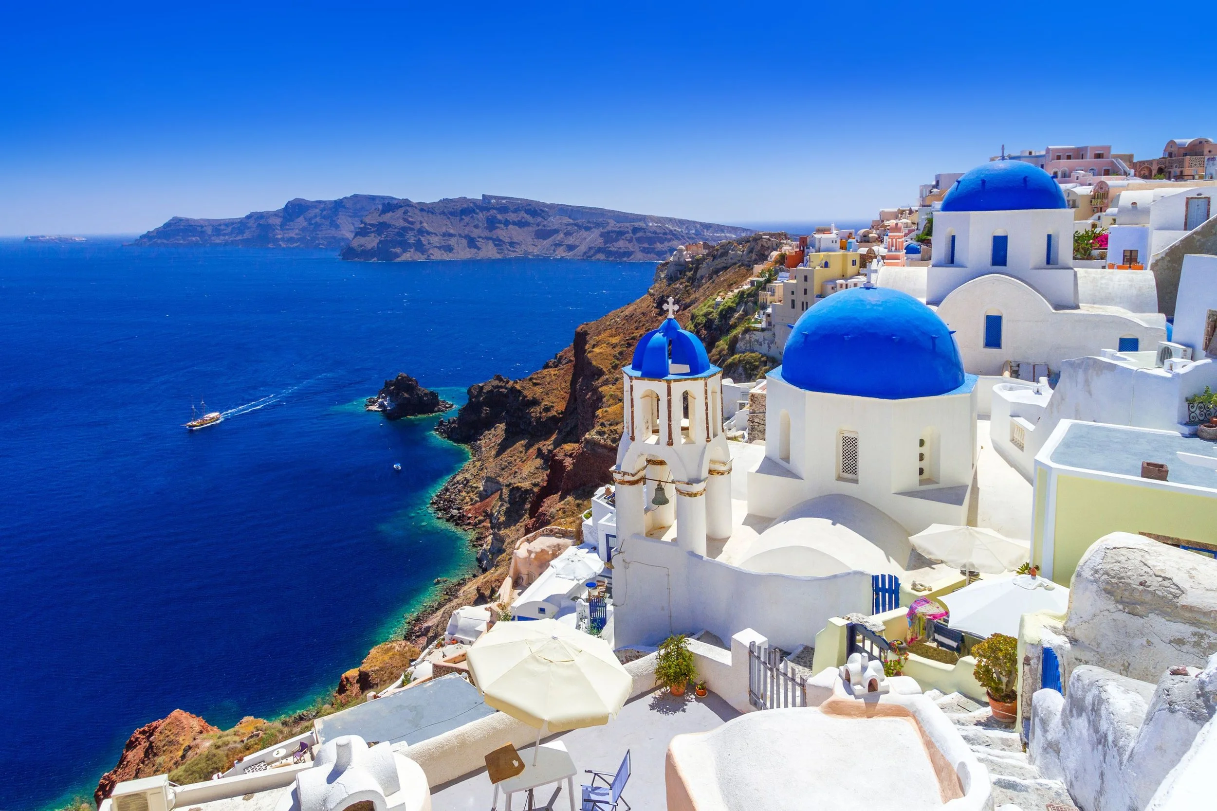 White buildings with blue domes on a cliffside overlooking the deep blue sea in Santorini, Greece, under a clear sky.