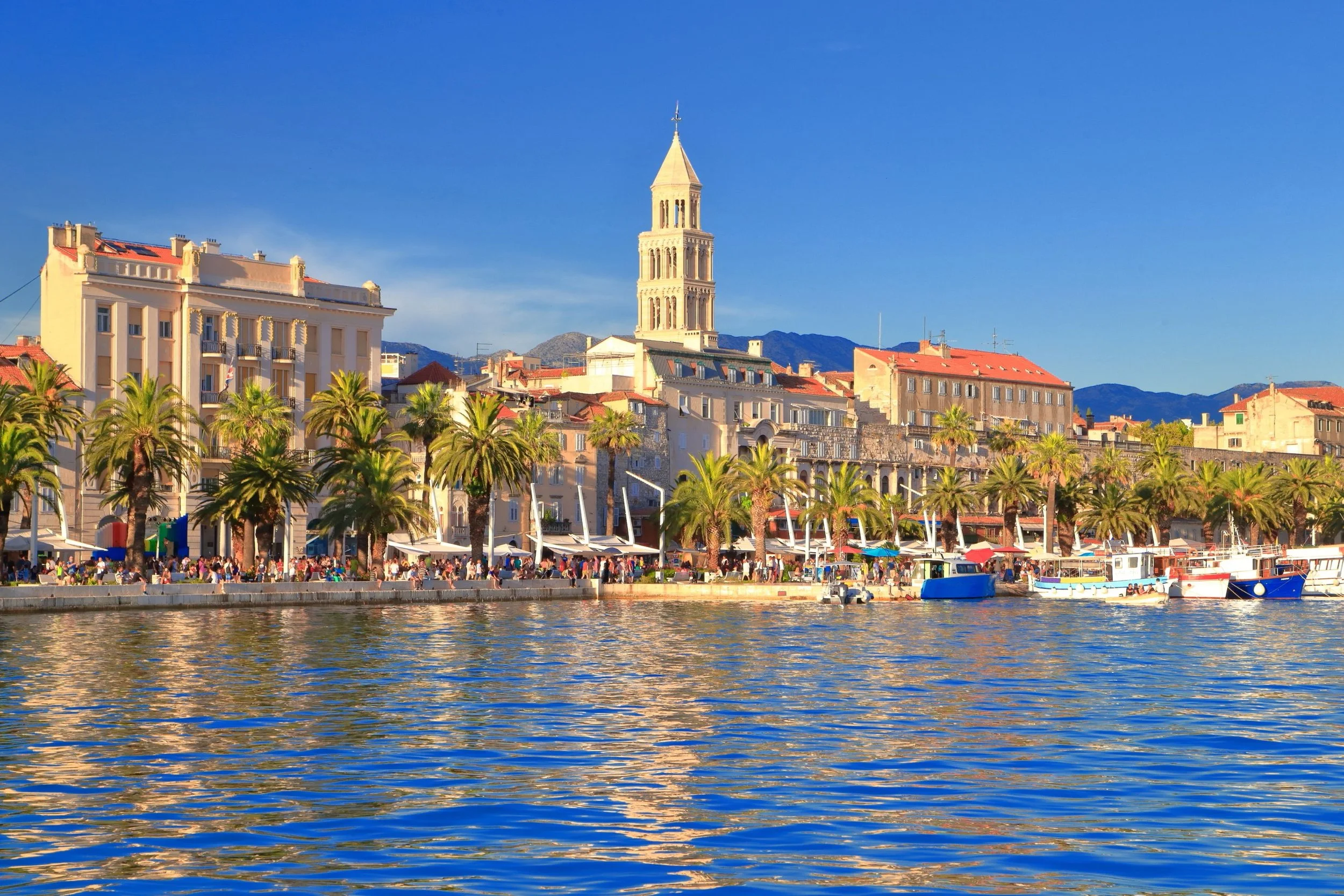 View of a waterfront city with colorful buildings, palm trees, boats on the water, and mountains in the background on a clear sunny day.