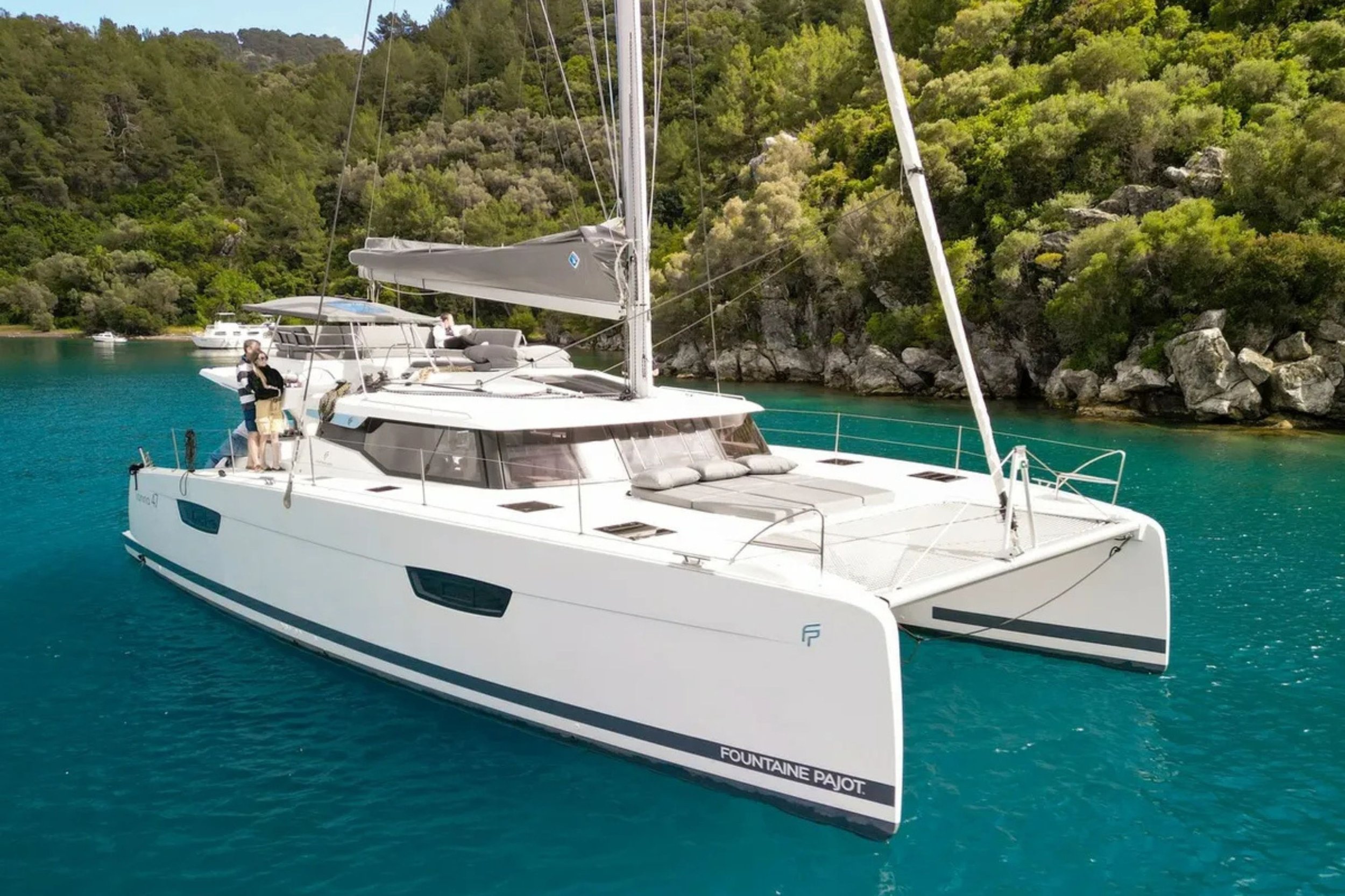 A white catamaran sailboat named Fountaine Pajot floating on turquoise water near a rocky, green hillside.