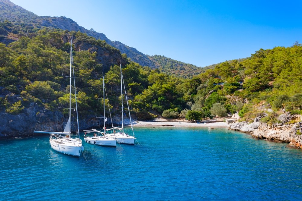 Two sailboats anchored in a calm, clear blue bay surrounded by green hills and rocky cliffs on a sunny day.