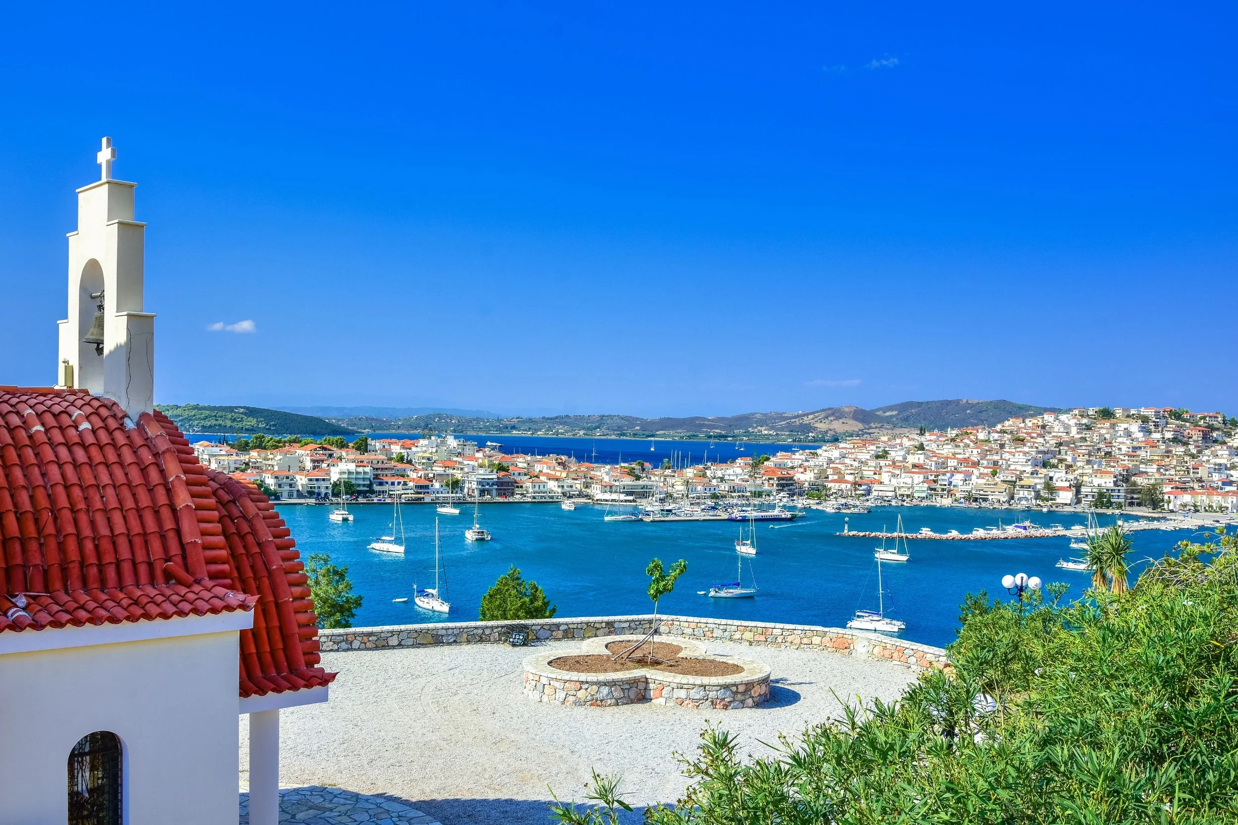 A coastal town with colorful buildings along a harbor filled with sailboats, viewed from a terrace with a small sculpture of a tree in a circular stone planter and a nearby white church with a bell tower.