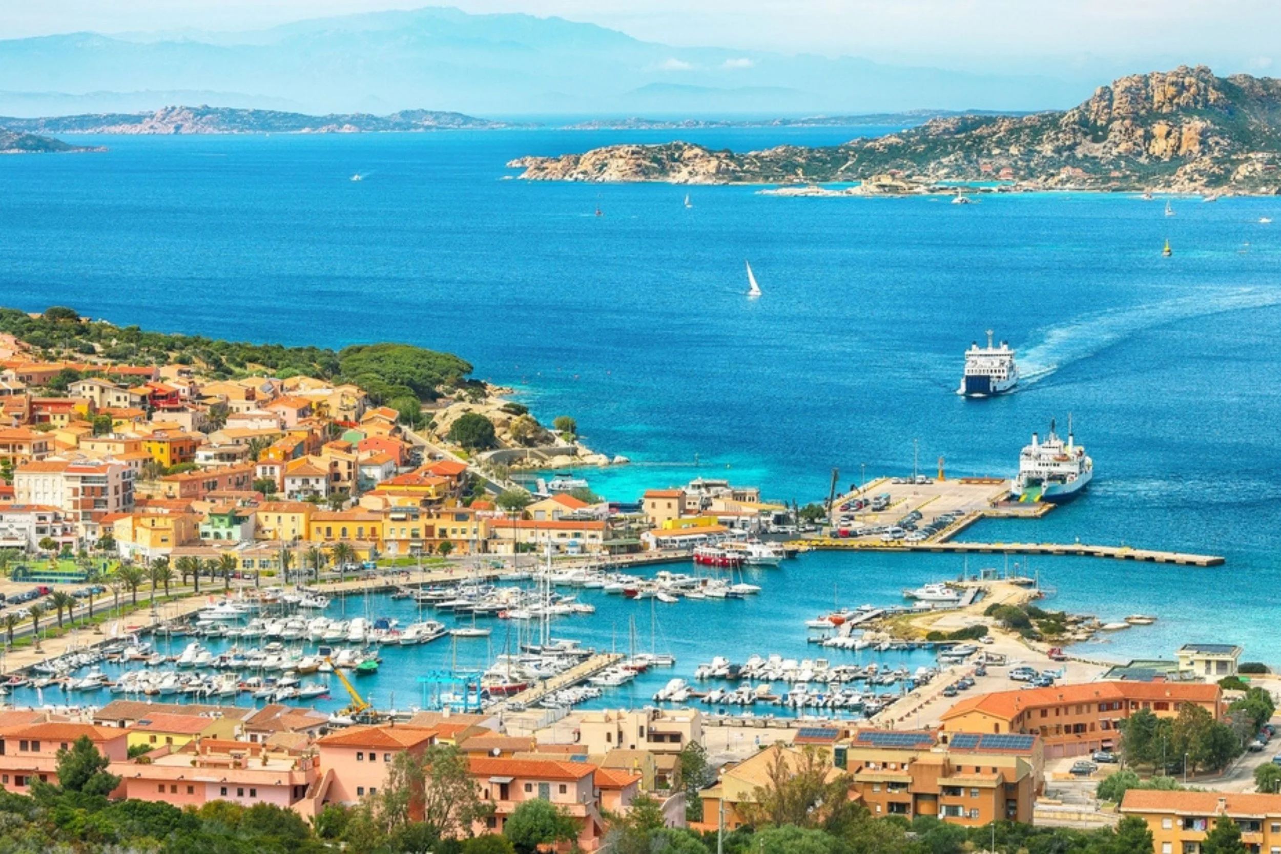 Aerial view of a harbor with boats, yachts, and ships, surrounded by a colorful town with houses and buildings along the coastline, and mountains in the distance across the blue water.