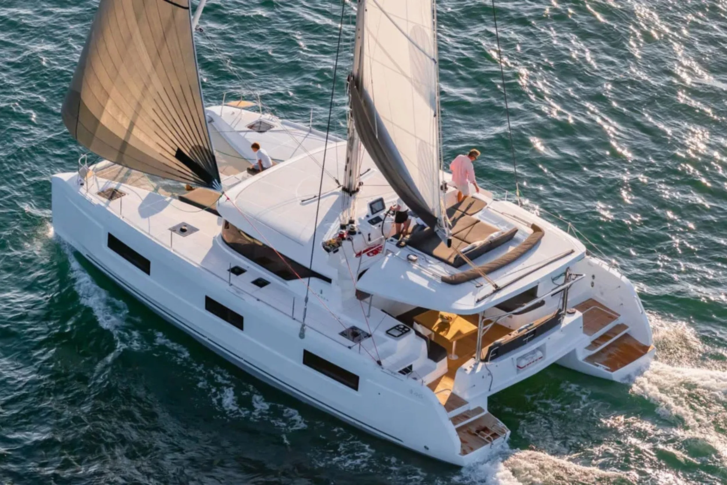 Aerial view of a white sailing yacht on the water with two people on deck, large sails, and seating areas.