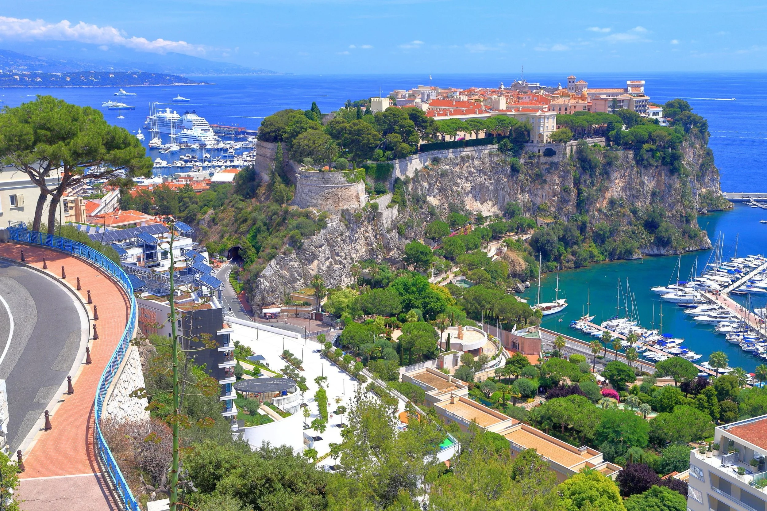 A coastal city with a marina filled with yachts, a fortress atop a hill, lush green trees, and Mediterranean-style buildings overlooking the blue sea under a partly cloudy sky.