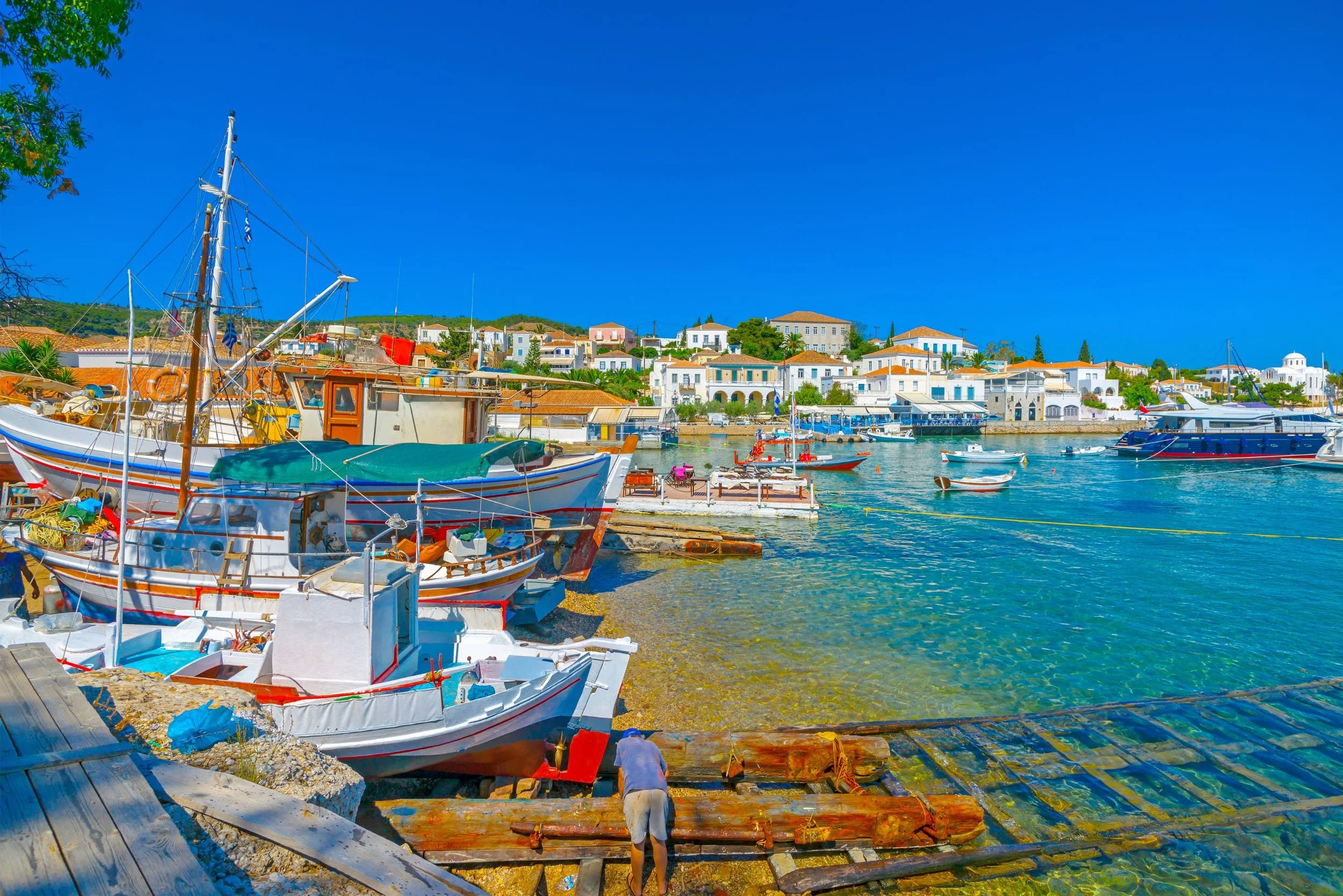 A colorful harbor with several boats docked along a waterfront, with a seaside town featuring white buildings and orange rooftops in the background under a clear blue sky.