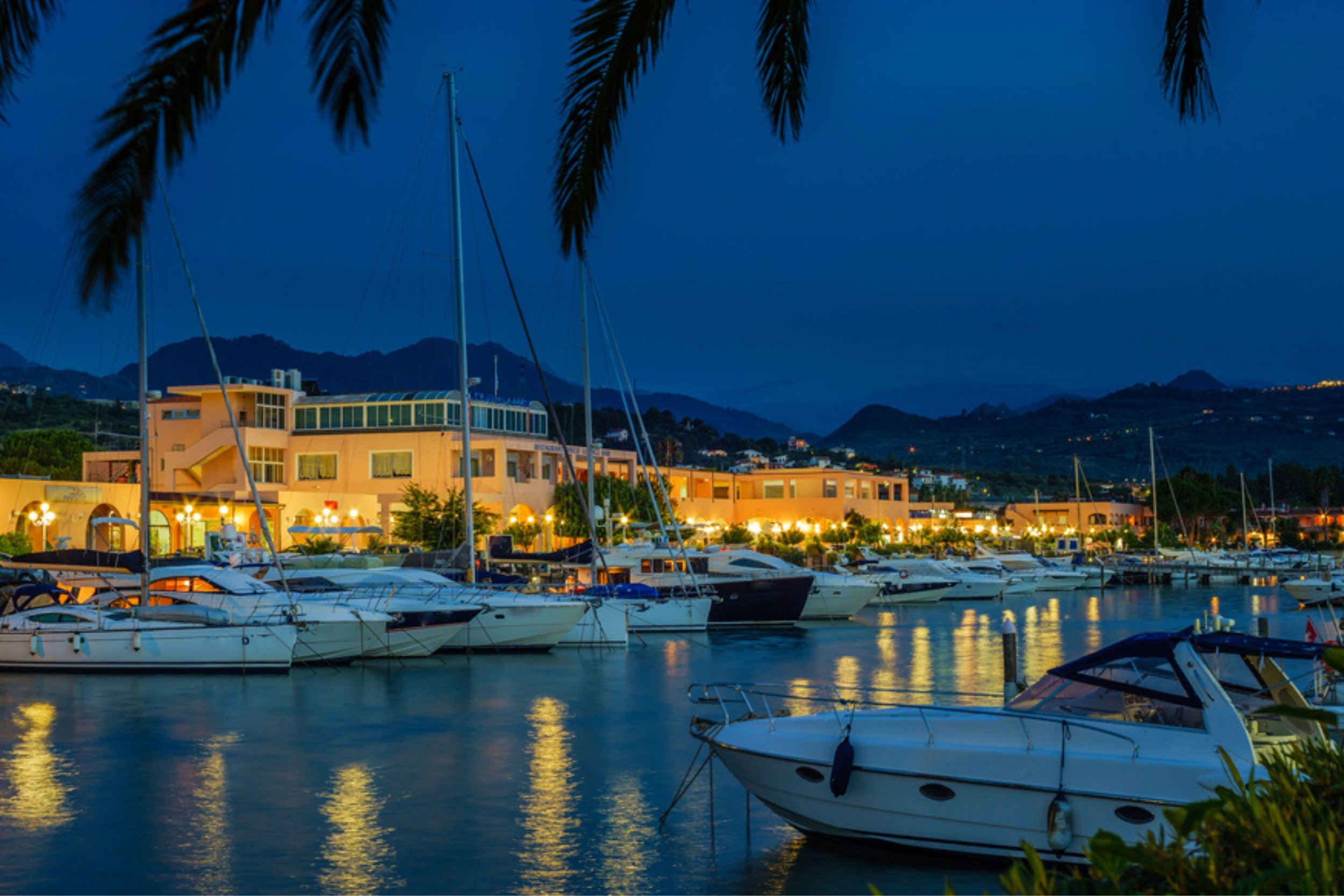 Nighttime view of a marina with docked sailboats and motorboats, illuminated by lights from nearby buildings, with mountains in the background and palm tree fronds in the foreground.