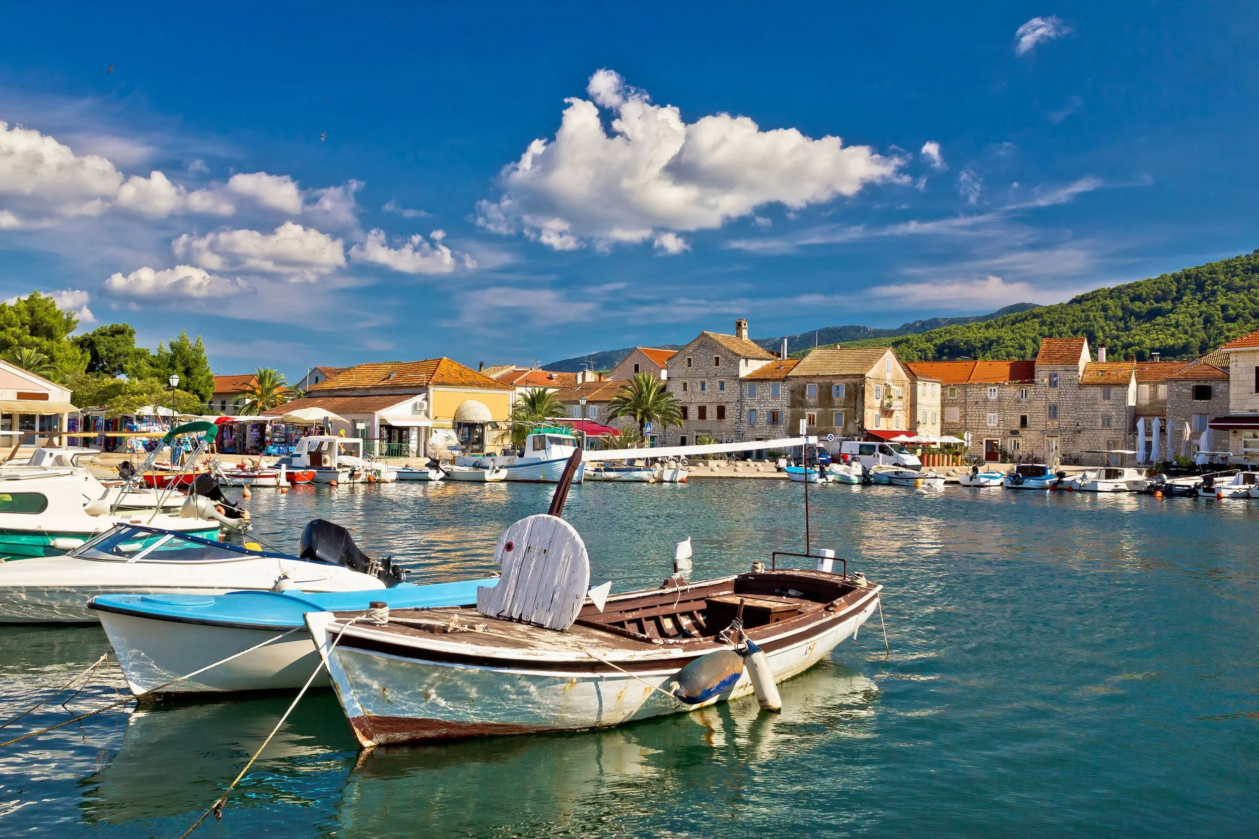 A scenic view of a small harbor with boats docked along the waterfront, colorful buildings with red-tiled roofs lining the shore, green hills in the background, and a partly cloudy sky overhead.