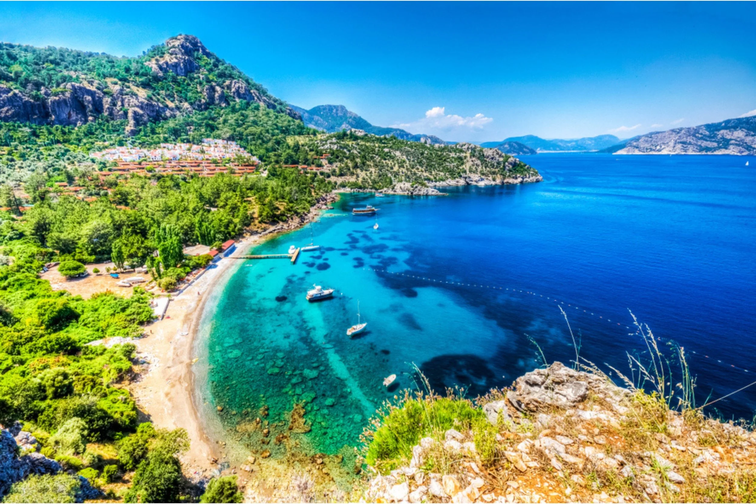 A scenic coastal landscape with a sandy beach, clear turquoise water, boats anchored near the shore, lush green trees, and rolling hills in the background under a bright blue sky.