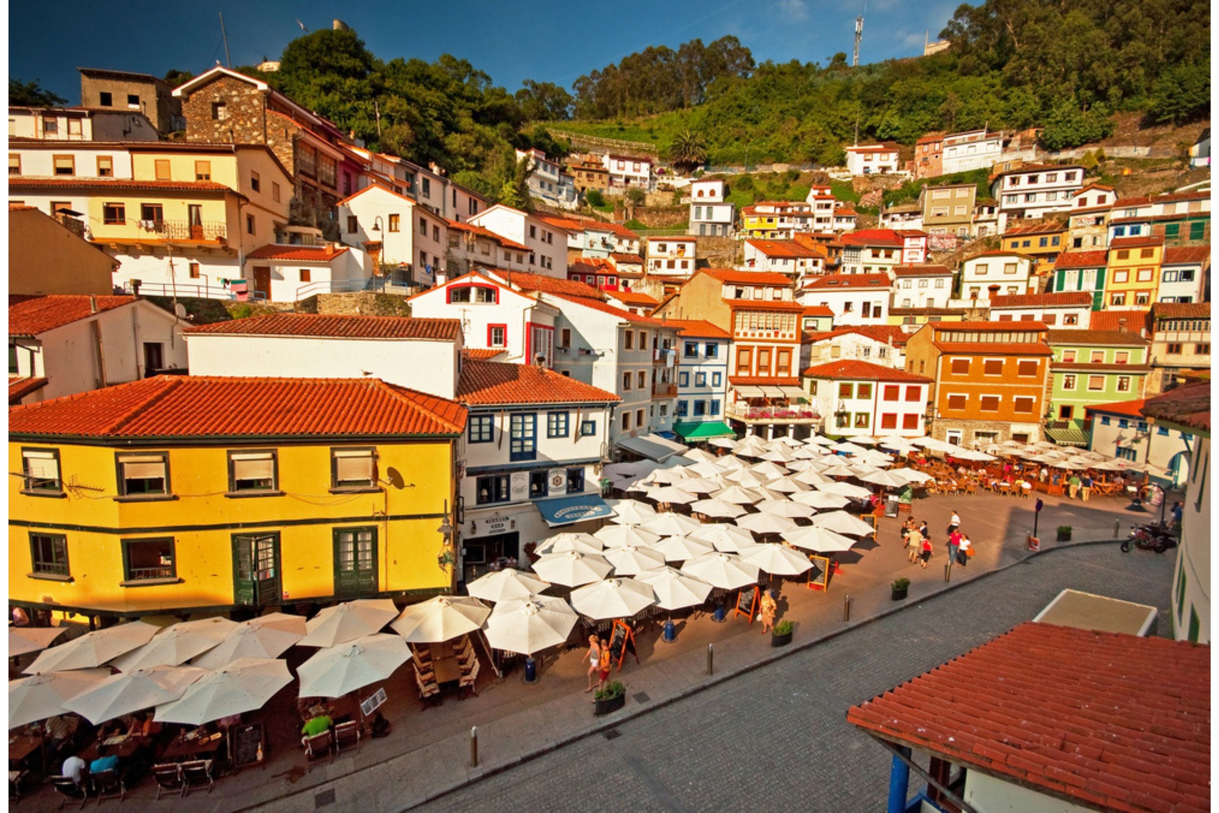 Colorful hillside town with white and multicolored buildings, a sunny outdoor café with white umbrellas, and people walking along a cobblestone street.