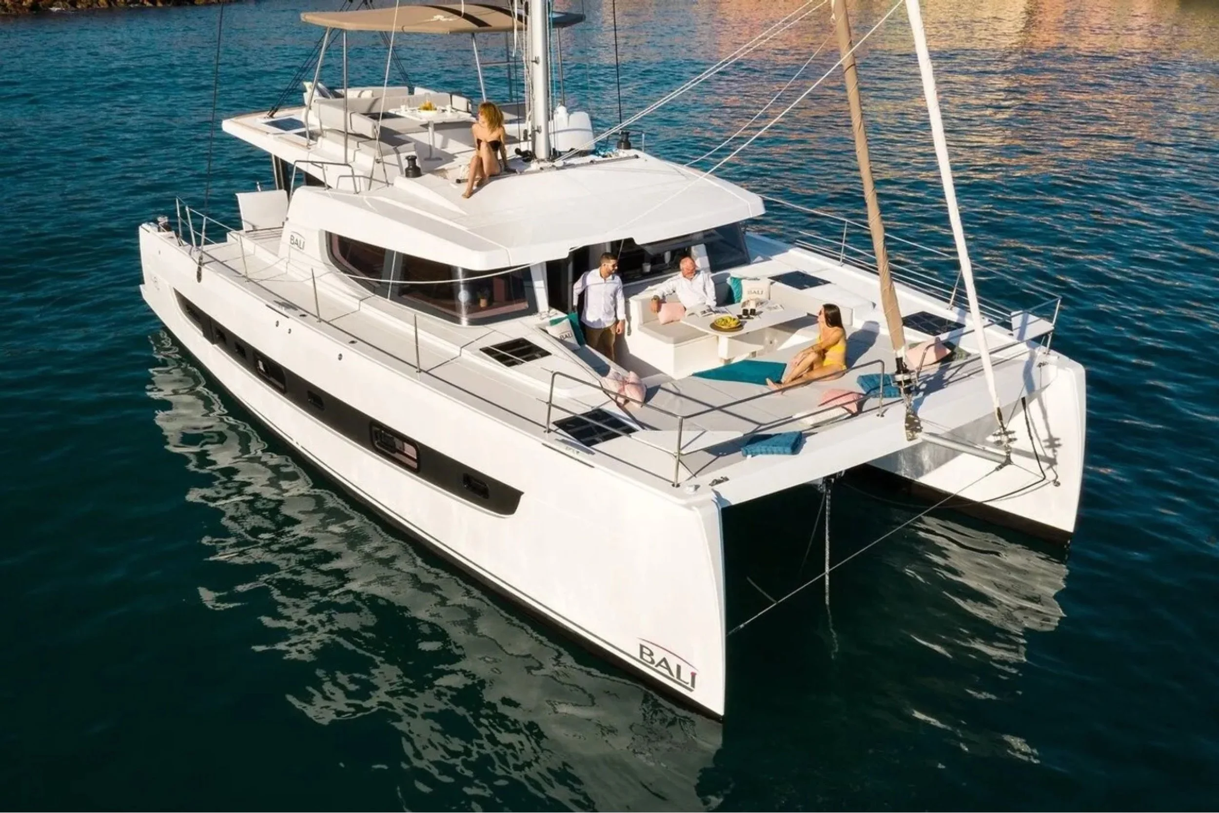 A white sailing yacht named Bali on the water with four people relaxing and socializing on the deck and a woman sitting on the upper deck.