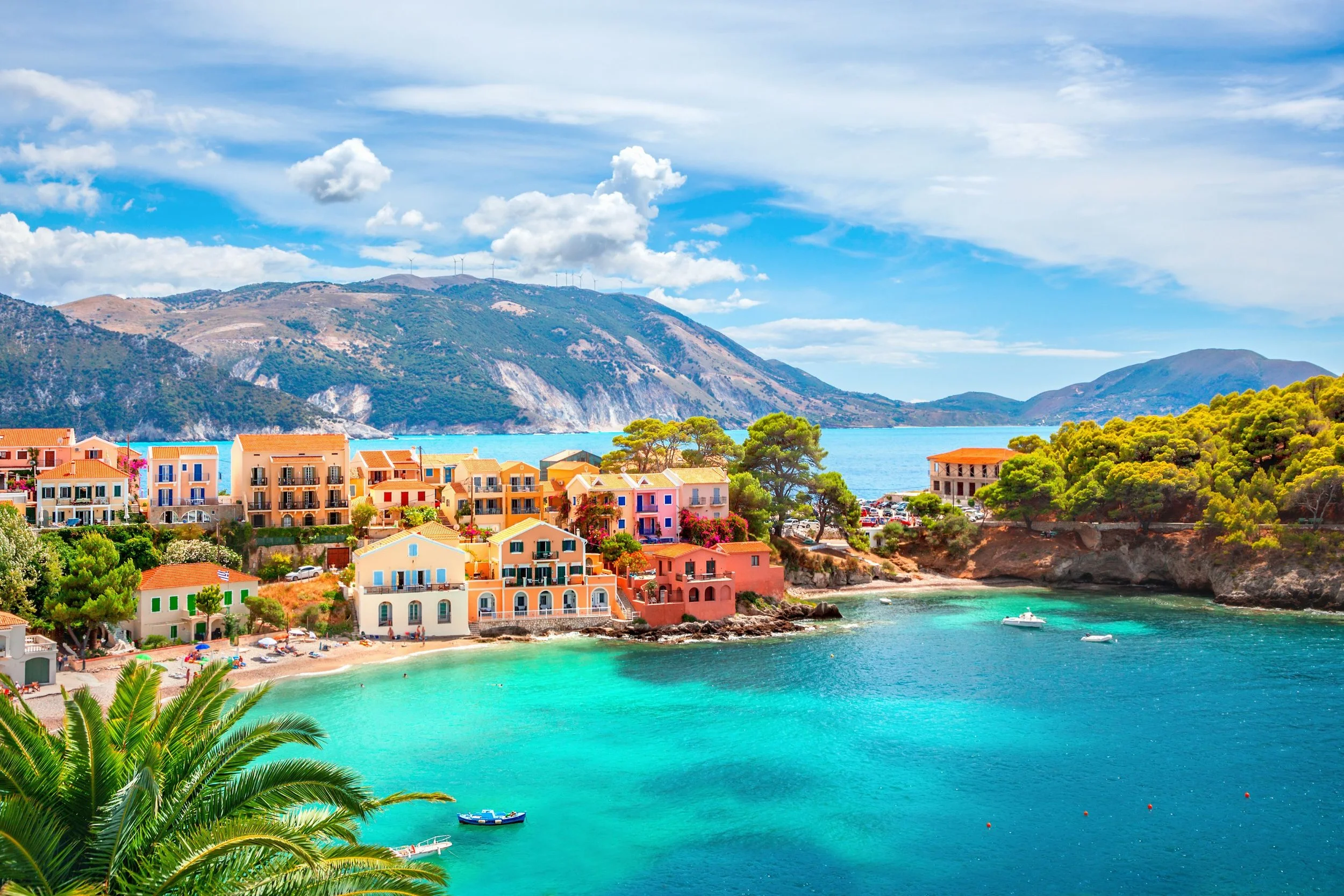 Colorful seaside village with pastel houses, turquoise water, boats, and lush greenery, with mountains in the background under a partly cloudy sky.