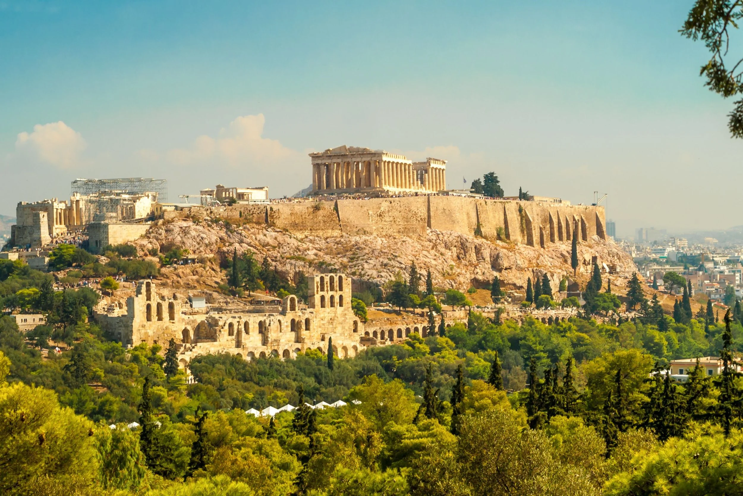 Panoramic view of the Acropolis in Athens, Greece, with the Parthenon atop the hill and ancient ruins in the foreground, surrounded by lush green trees.