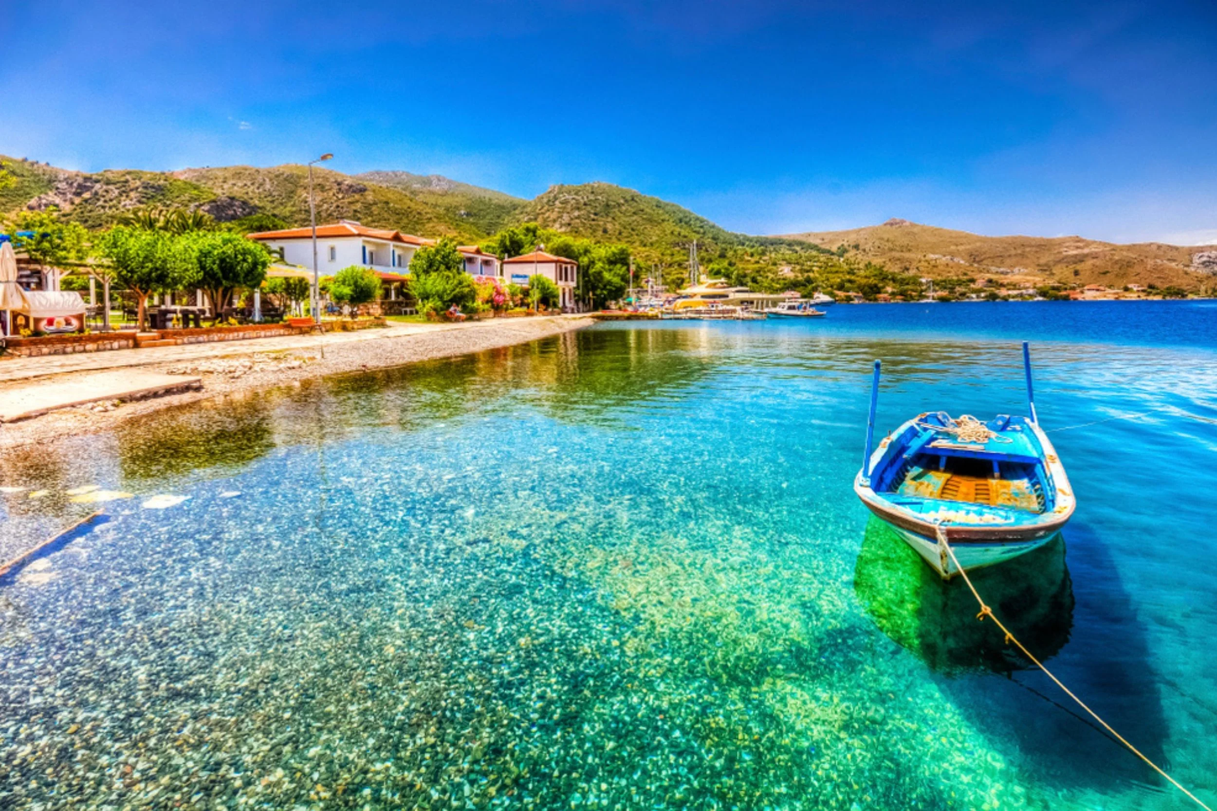 Small boat anchored near the rocky shoreline of a clear, turquoise bay with white houses, greenery, and mountains in the background on a sunny day.