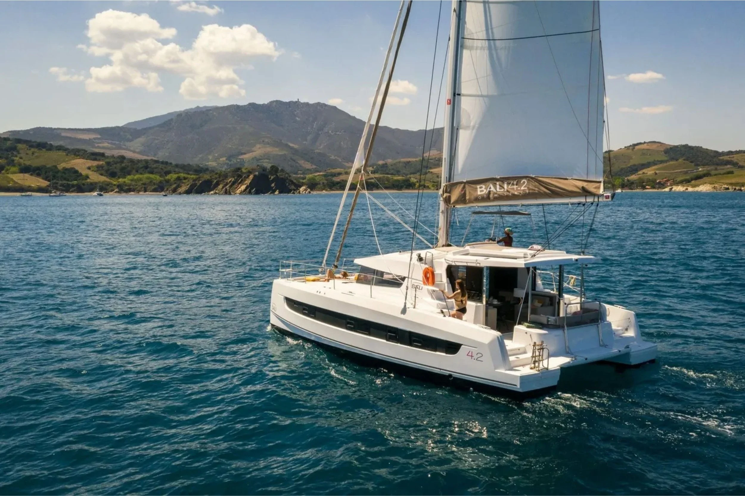 A white sailing yacht on blue water with a hilly landscape and mountains in the background, featuring a person onboard near the helm and another closer to the bow.