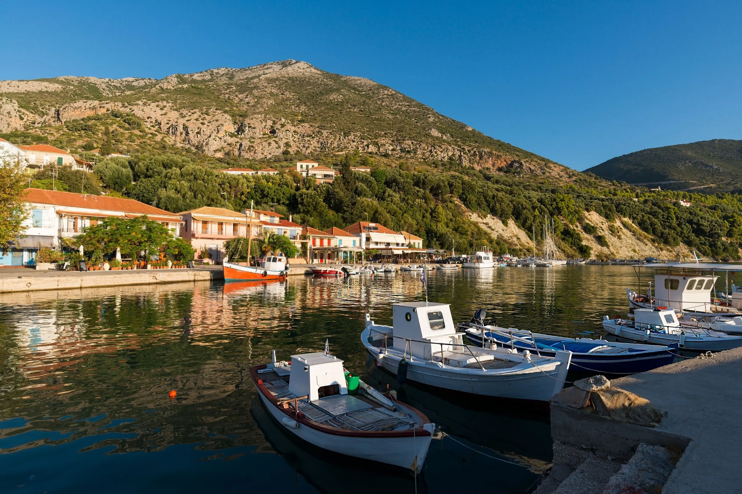 A peaceful seaside village with boats docked along a calm harbor, colorful houses on the shoreline, lush green hills and rocky mountains in the background, under a clear blue sky.