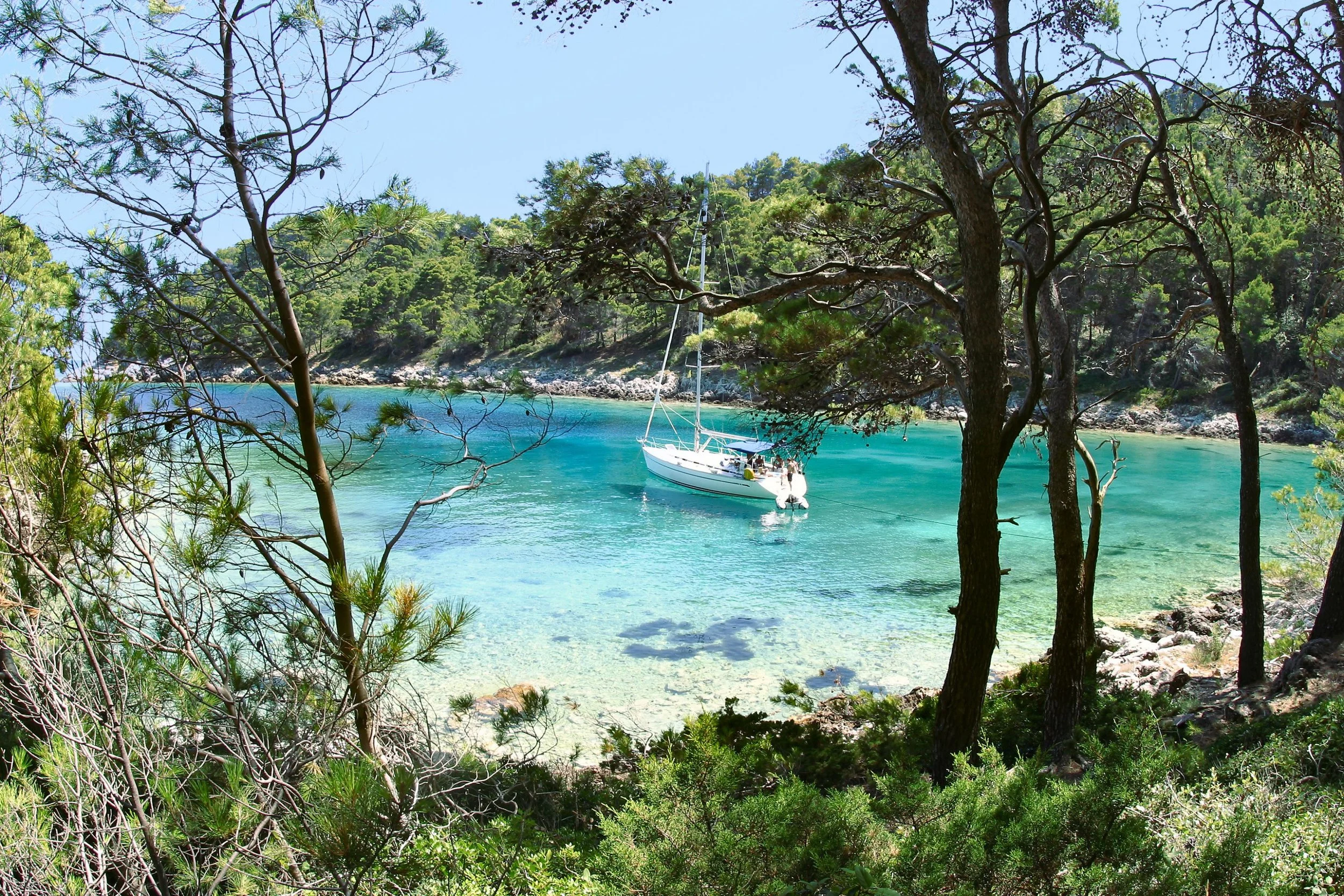 A scenic view of a turquoise bay with a white sailboat anchored near the shore. The foreground has trees and green bushes, and the background features a lush, forested hillside under a clear blue sky.