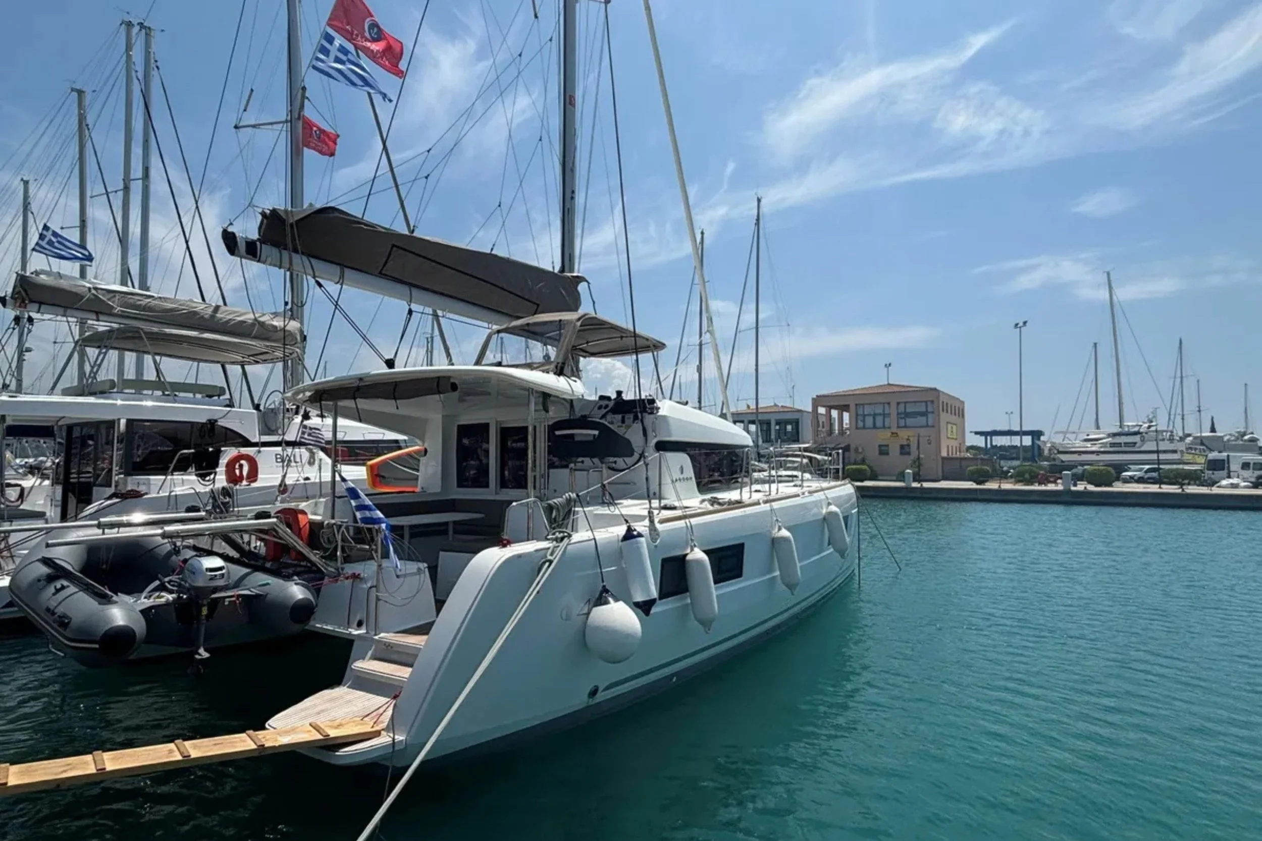 A marina with sailboats and yachts docked in the water under a partly cloudy sky, with buildings, a walkway, and parked cars visible in the background.