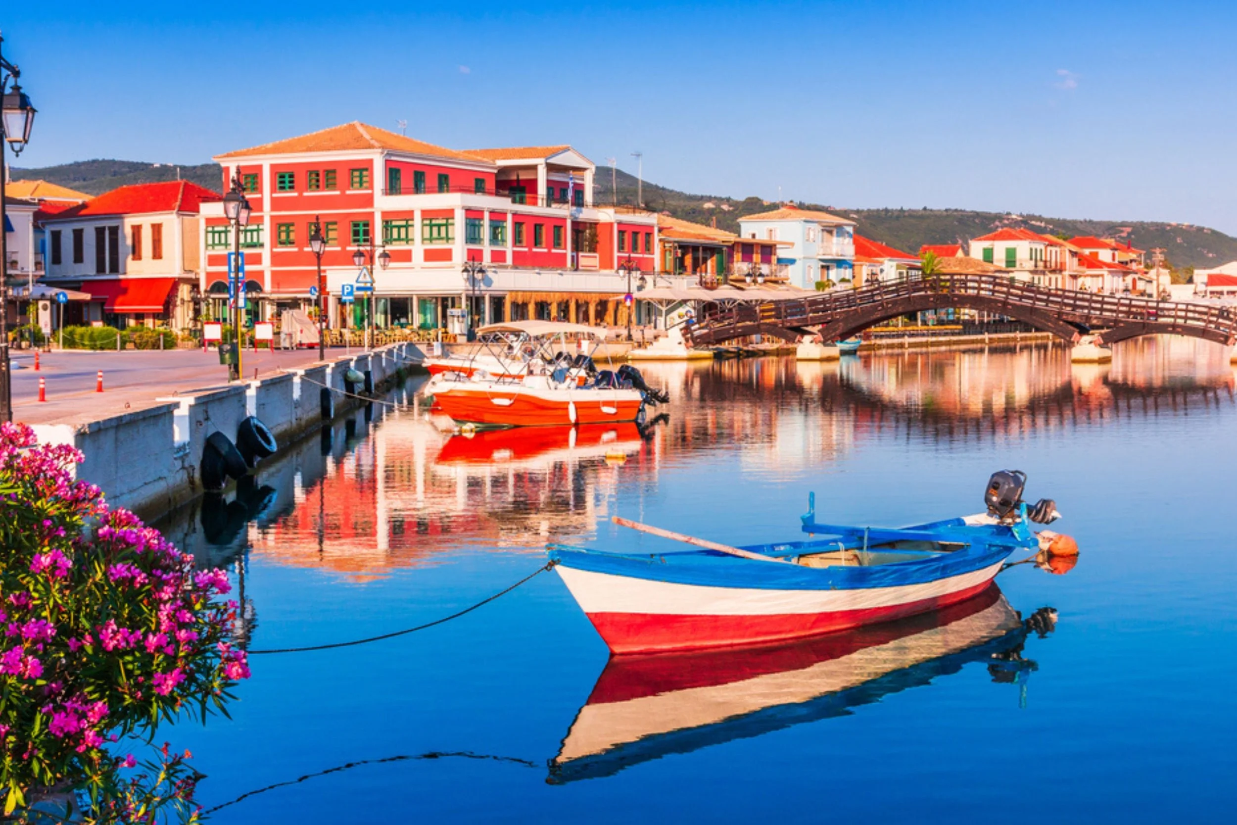 Colorful boats docked along a peaceful waterfront with vibrant buildings, a wooden bridge, and flowering plants reflected in calm water.