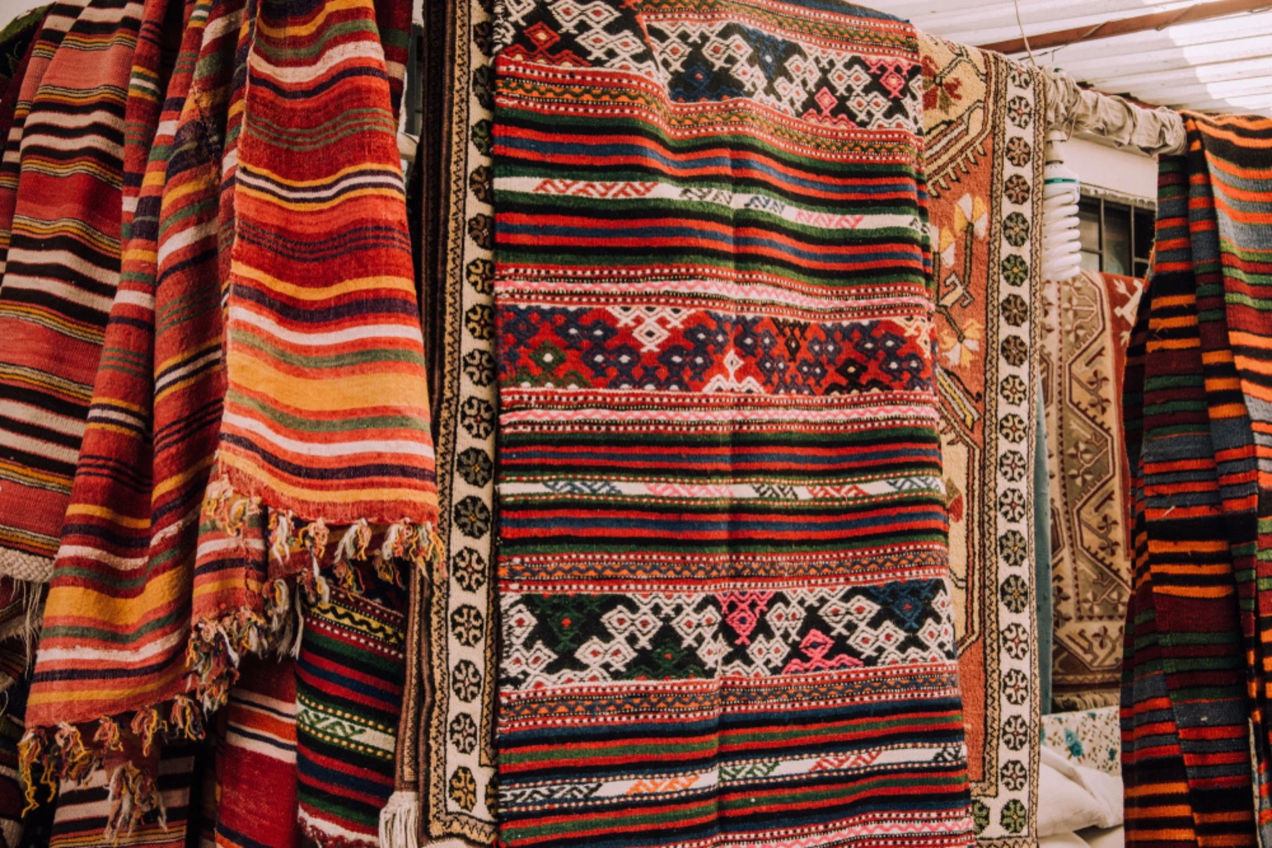 Display of colorful traditional woven rugs with intricate patterns and fringed edges at a market or store.