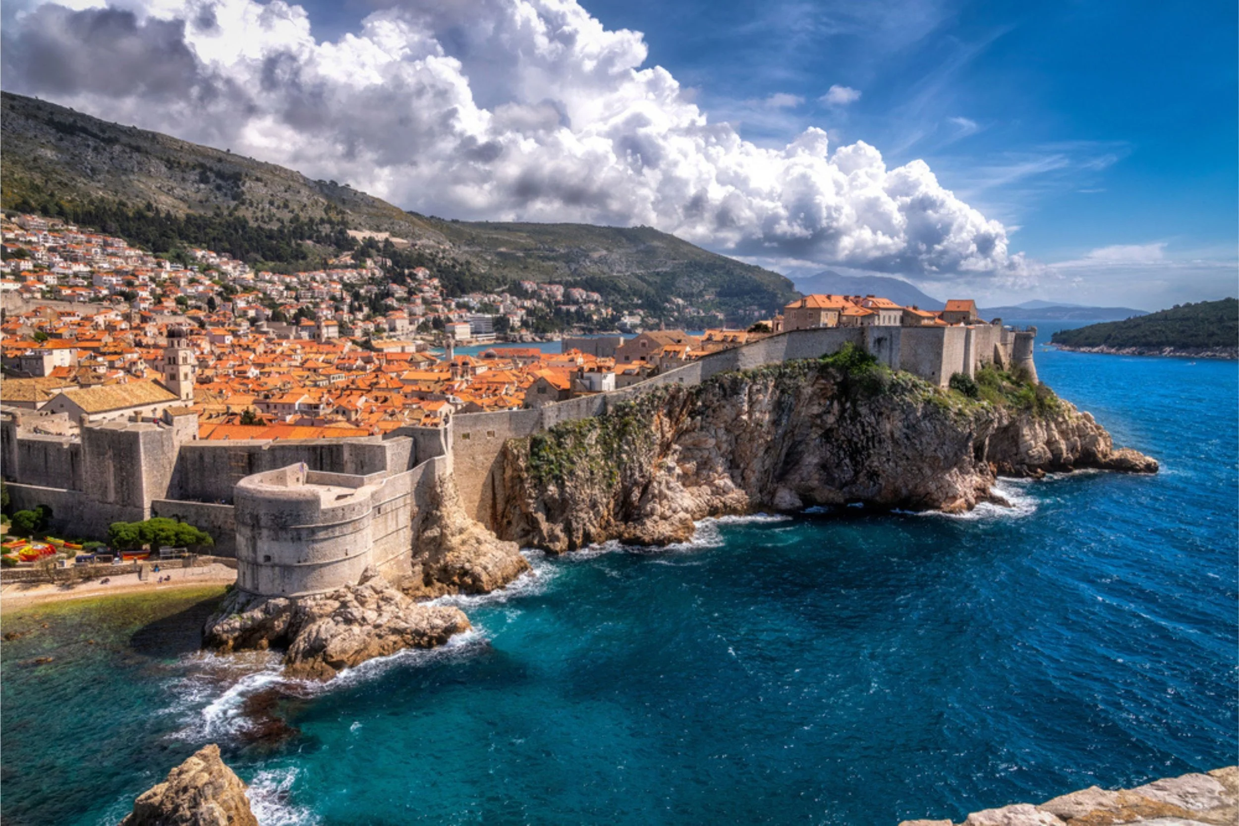 A coastal cityscape with a fortress wall overlooking the ocean, orange rooftops, a hillside filled with houses, mountains in the background, partly cloudy sky.
