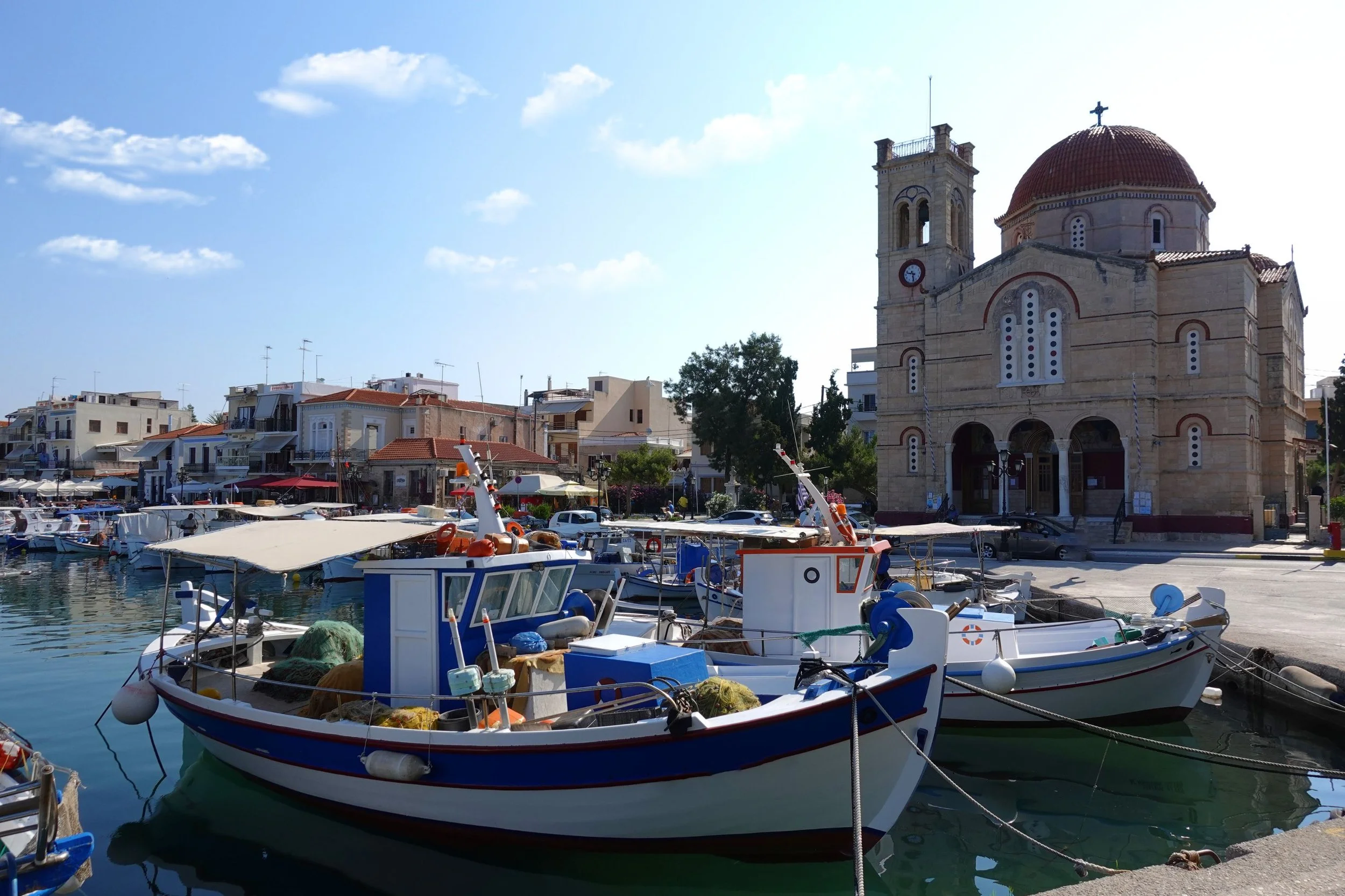 A harbor with several boats docked, a church with a red dome and a clock tower in the background, blue sky with clouds.