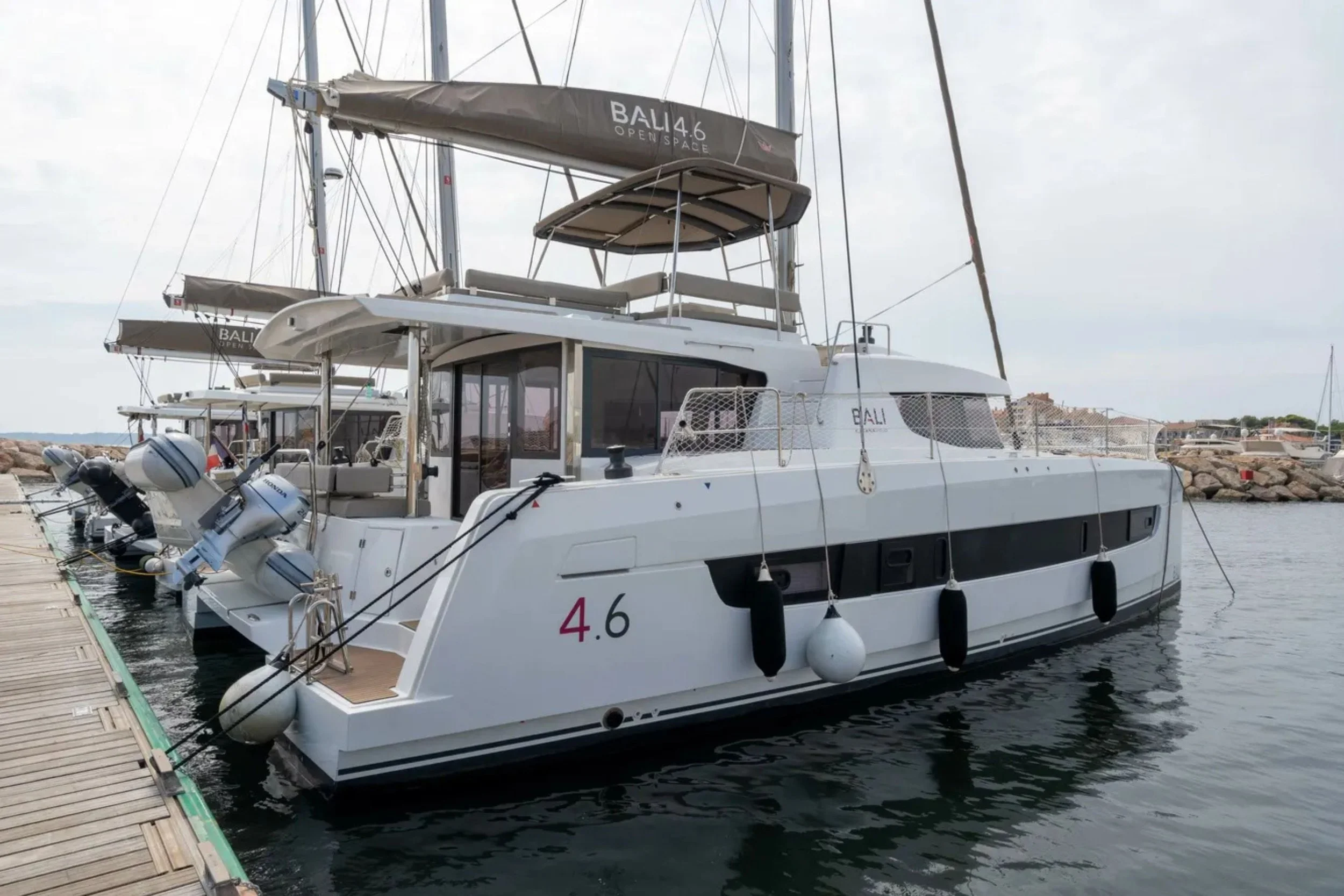 A white luxury yacht docked at a pier, with several similar yachts in the background, and rocks along the shoreline.